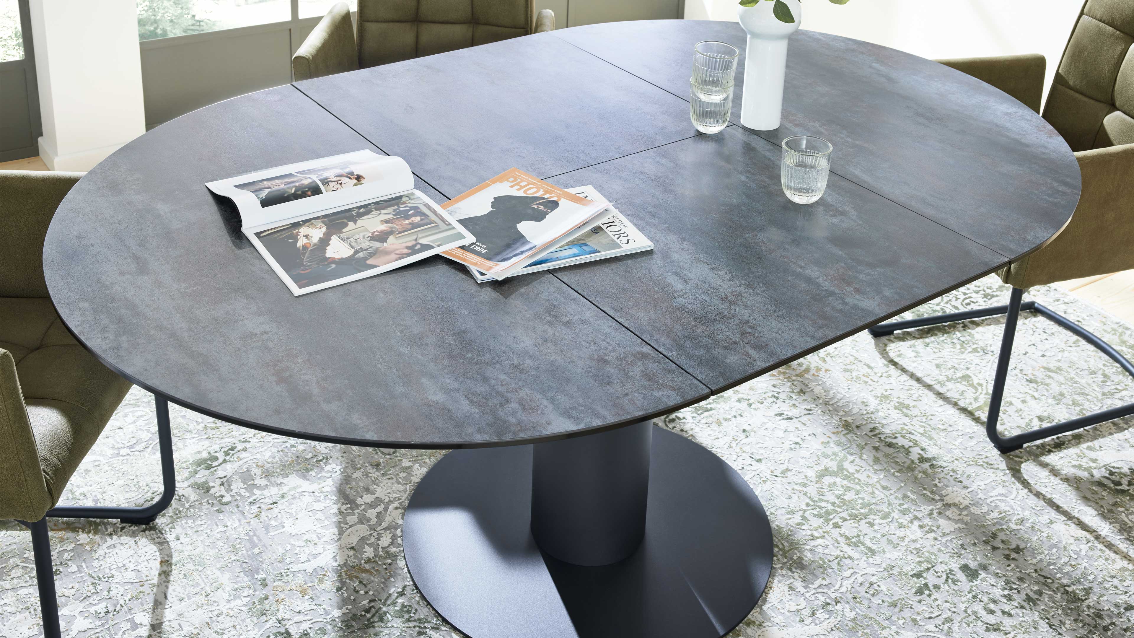 Ovaler dunkelgrauer Esstisch mit zentraler runder Säulenbasis, darauf Magazine, zwei Wassergläser und weiße Vase, umgeben von Stoffstühlen.