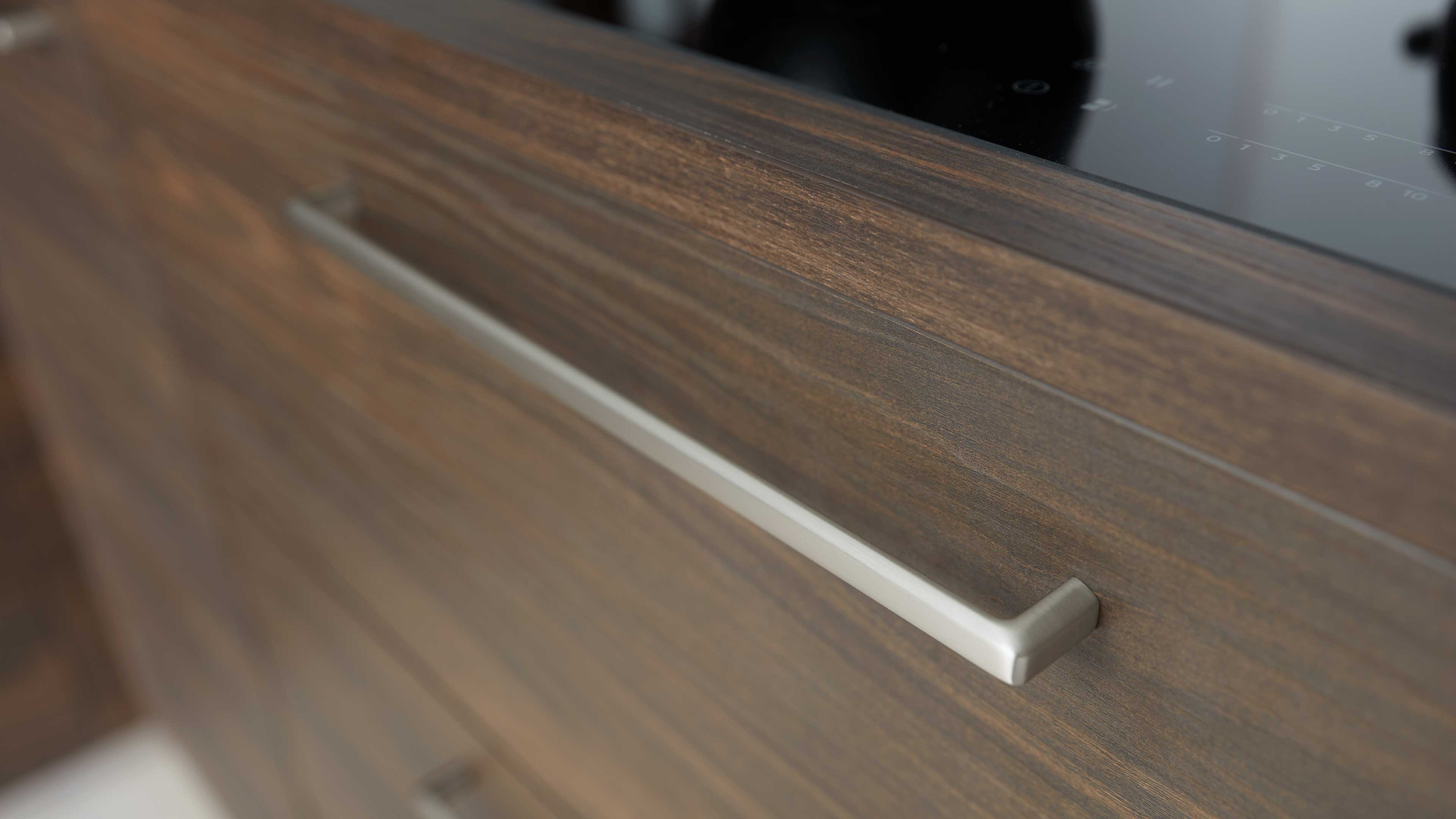 Front einer braunen Schublade mit sichtbarer Holzmaserung, langem mattierten Metallgriff und schwarzer Glasplatte oben.