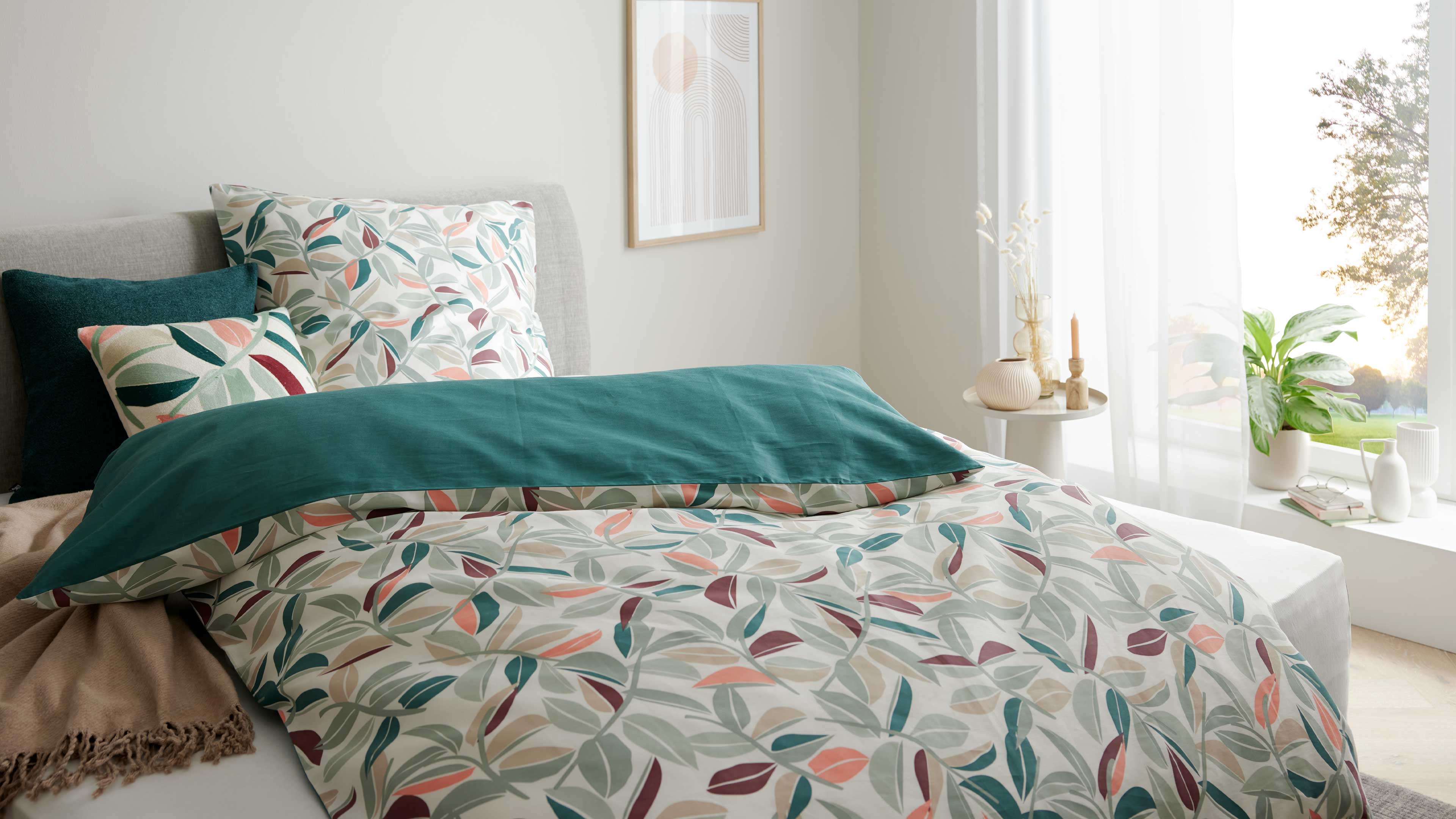 Bett mit grauem Kopfteil, Blattmuster-Bettbezug in Grün/Koralle, türkisfarbener Überwurf und Kissen, runder Nachttisch mit Vasen vor Fenster.