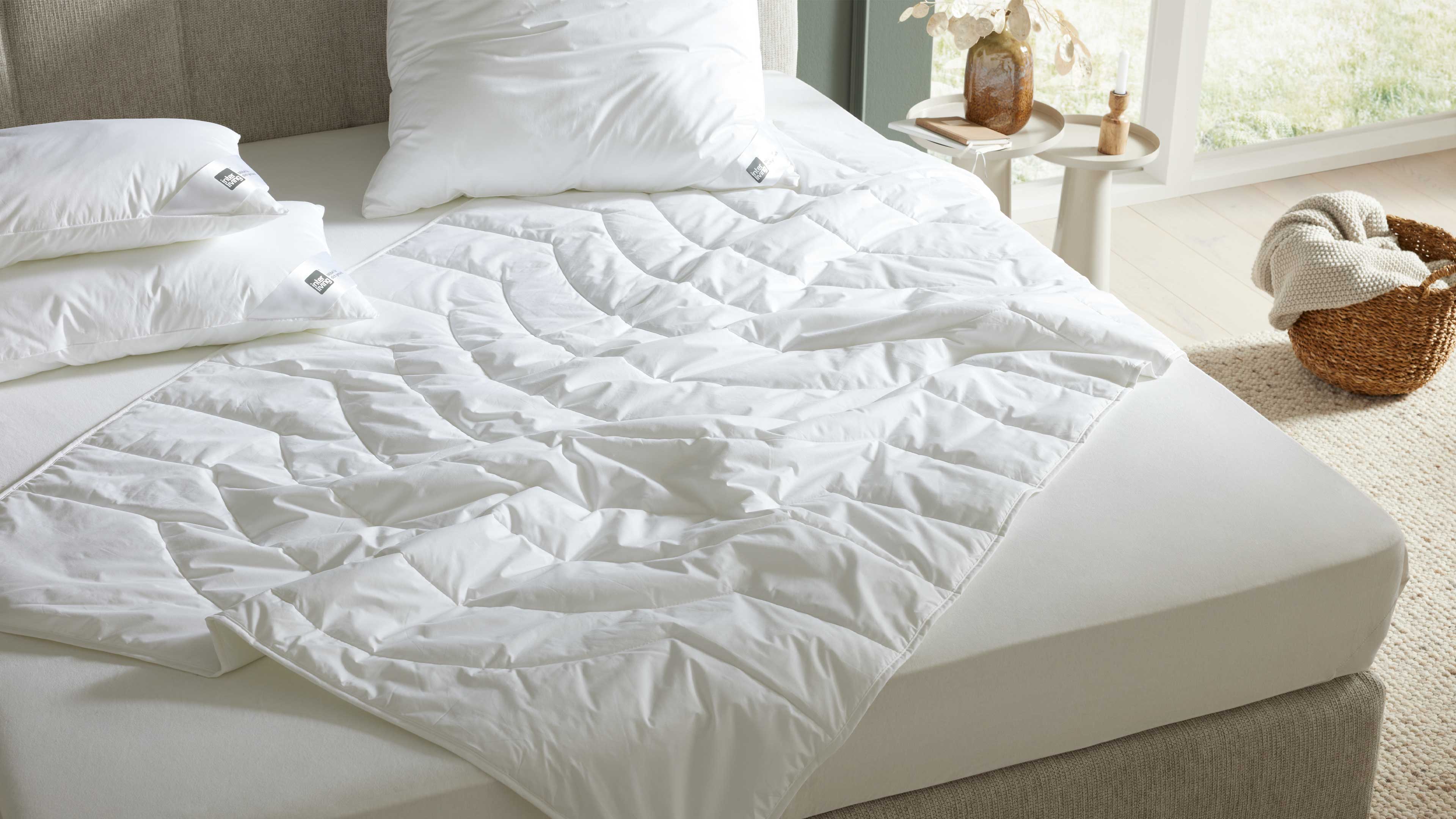 Weiße gesteppte Bettauflage liegt locker auf einer Matratze mit weißen Kissen; daneben Beistelltisch und Korb mit Decke.