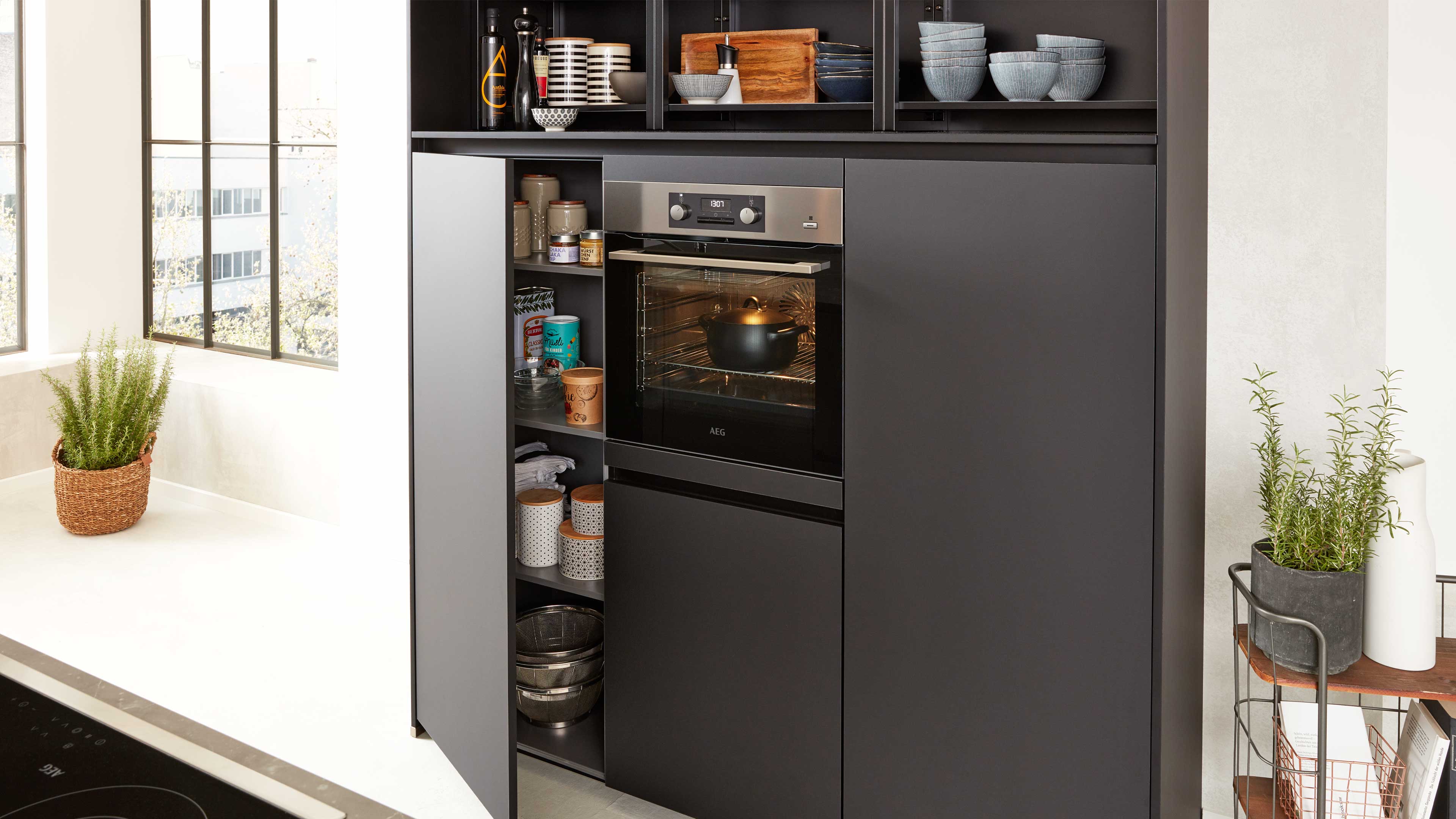 Dunkler Küchenhochschrank mit eingebautem Backofen und offenem Seitenfach, gefüllt mit Dosen, Schüsseln und Töpfen, daneben Pflanzen.