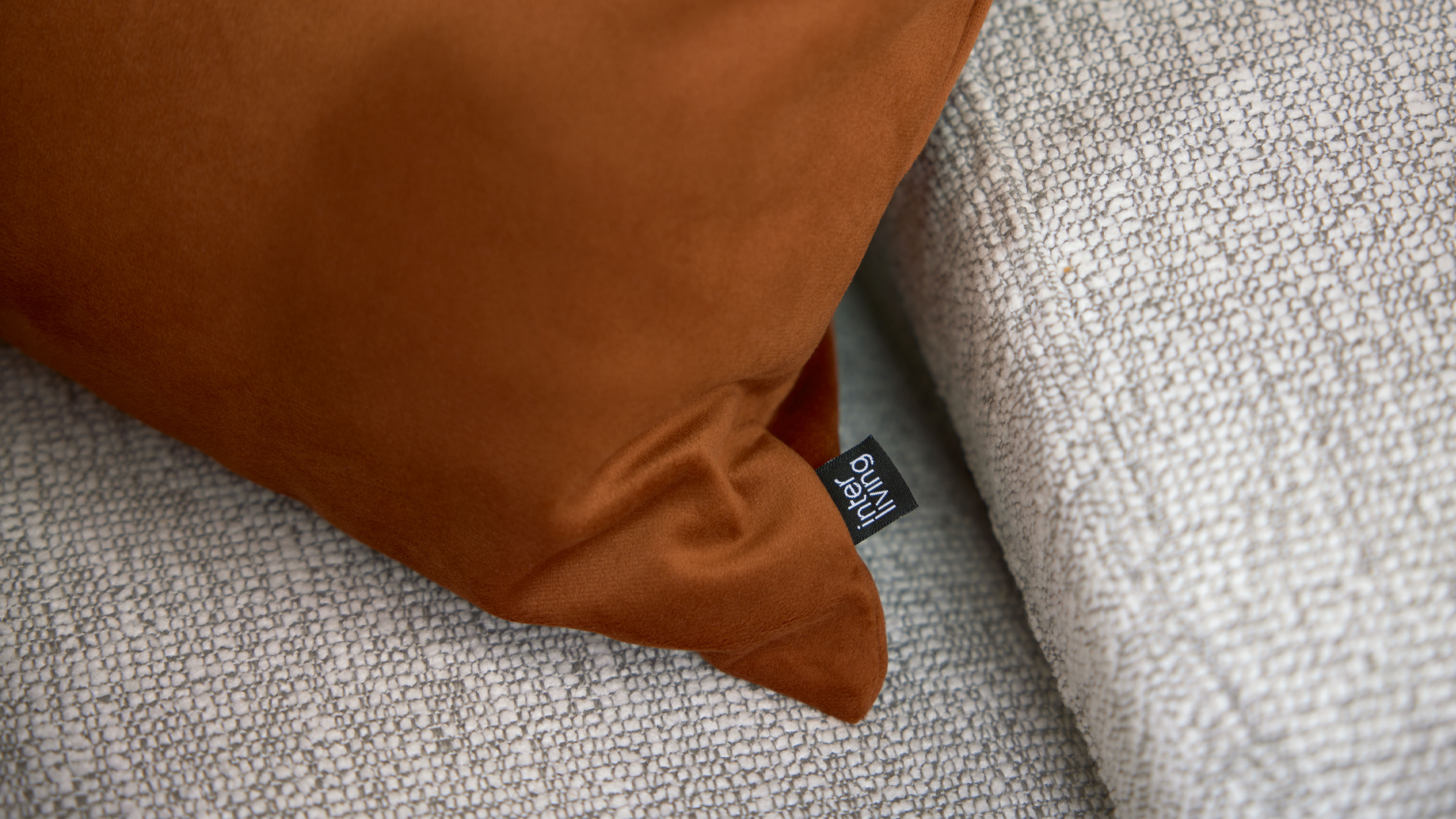 Rostbraunes Kissen liegt auf hellgrauem, grob gewebtem Sofa, Ecke mit kleiner Falte und sichtbarem schwarzem Etikett.