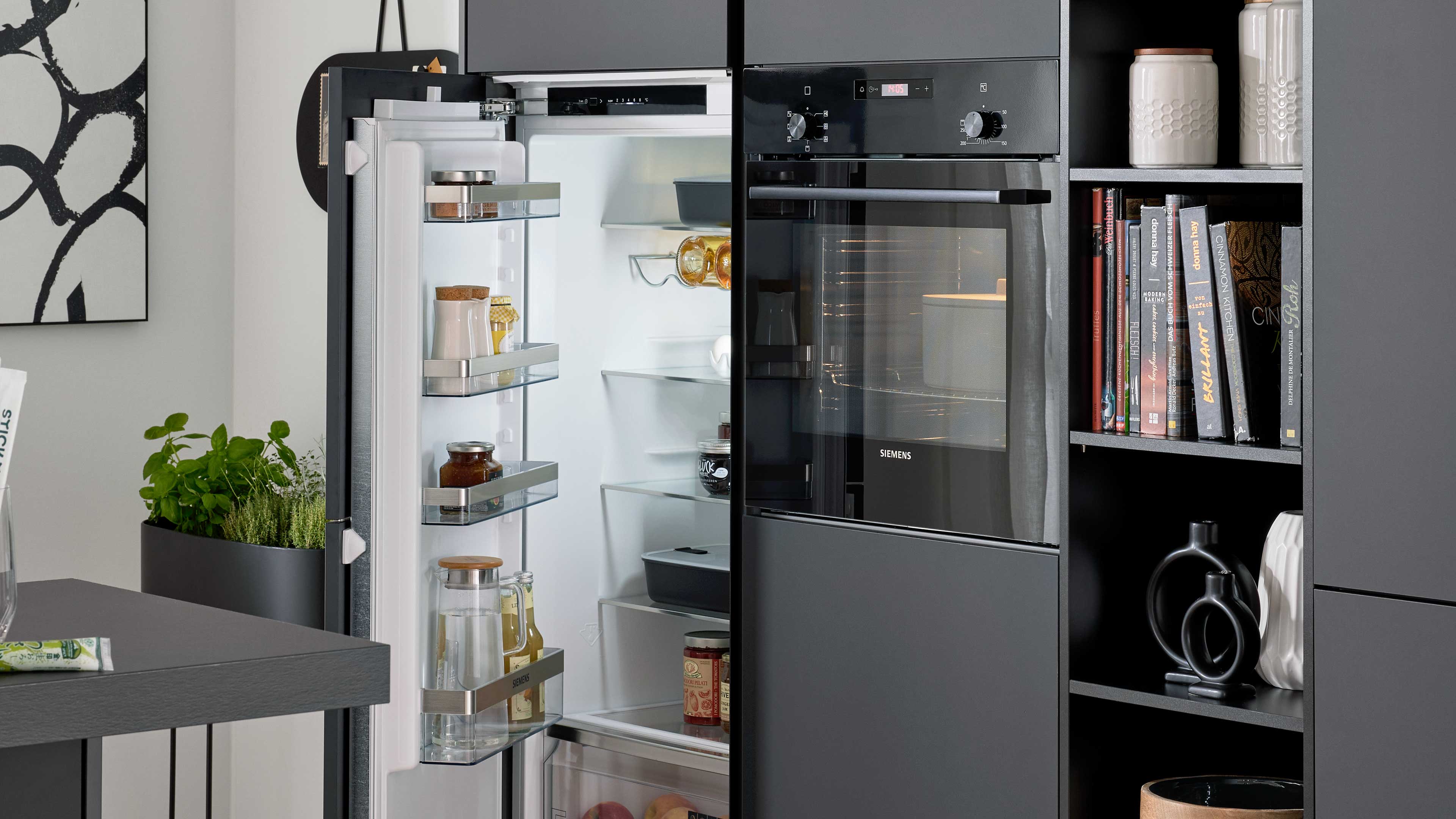 Geöffneter Einbaukühlschrank in dunkelgrauer Küchenzeile neben einem schwarzen Backofen, rechts offene Regalfächer mit Büchern und Dekor.