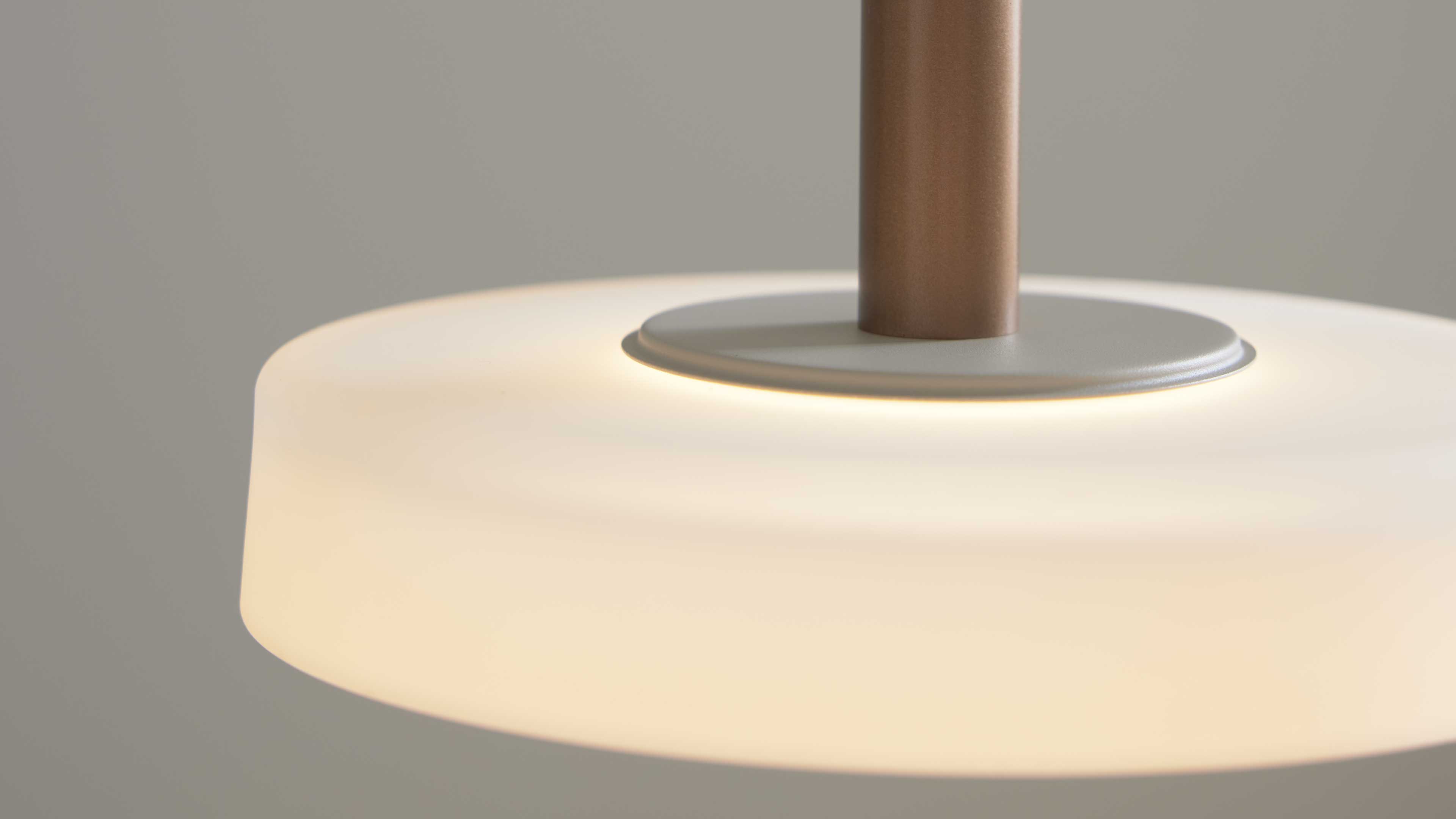 Pendelleuchte mit rundem opalweißen Glasschirm, warm leuchtender Oberfläche und zylindrischem bronzefarbenem Stab.