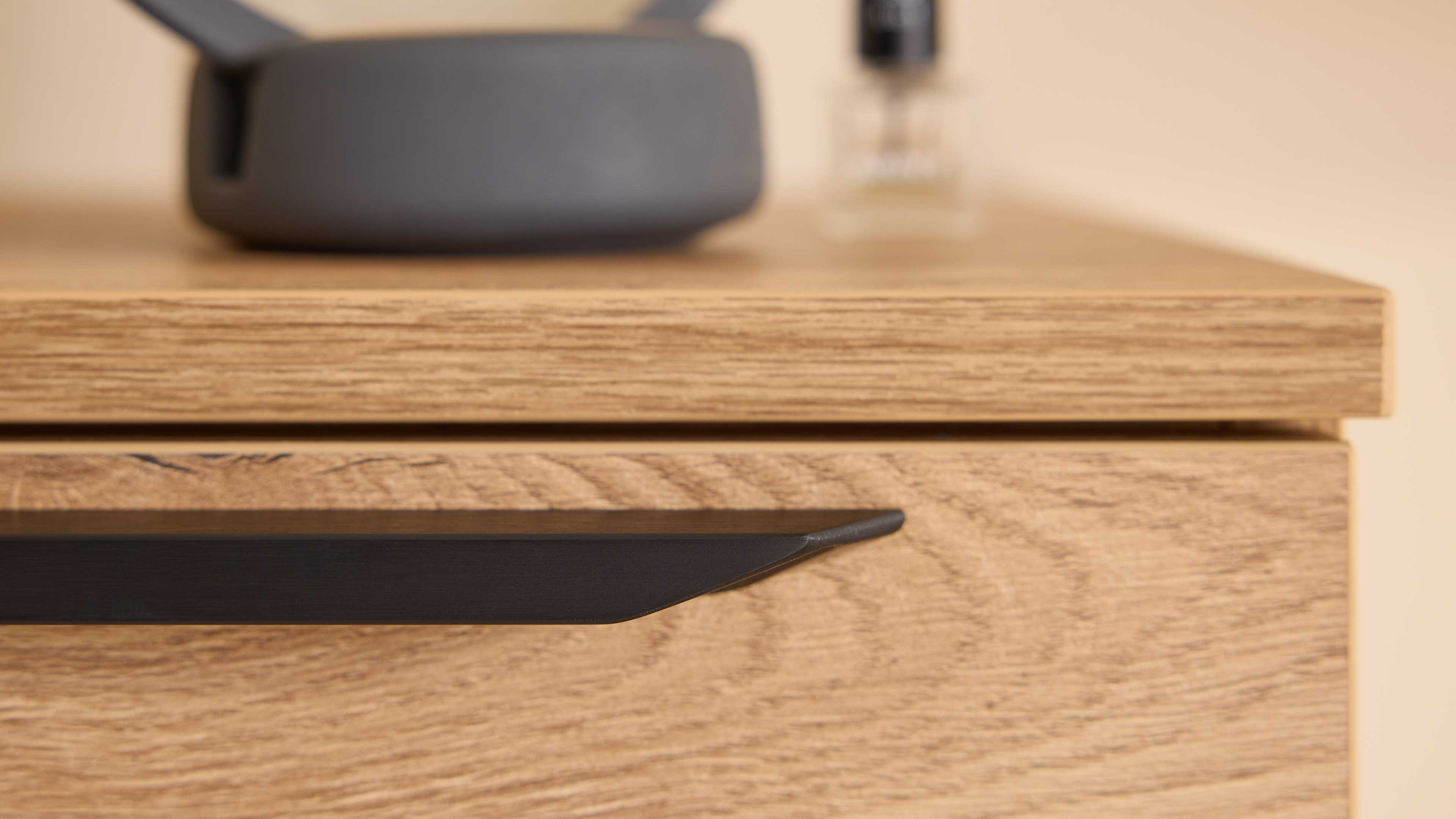 Helle Holz-Schublade mit sichtbarer Holzmaserung und flachem schwarzem Metallgriff, auf der Oberkante unscharfe Gegenstände.