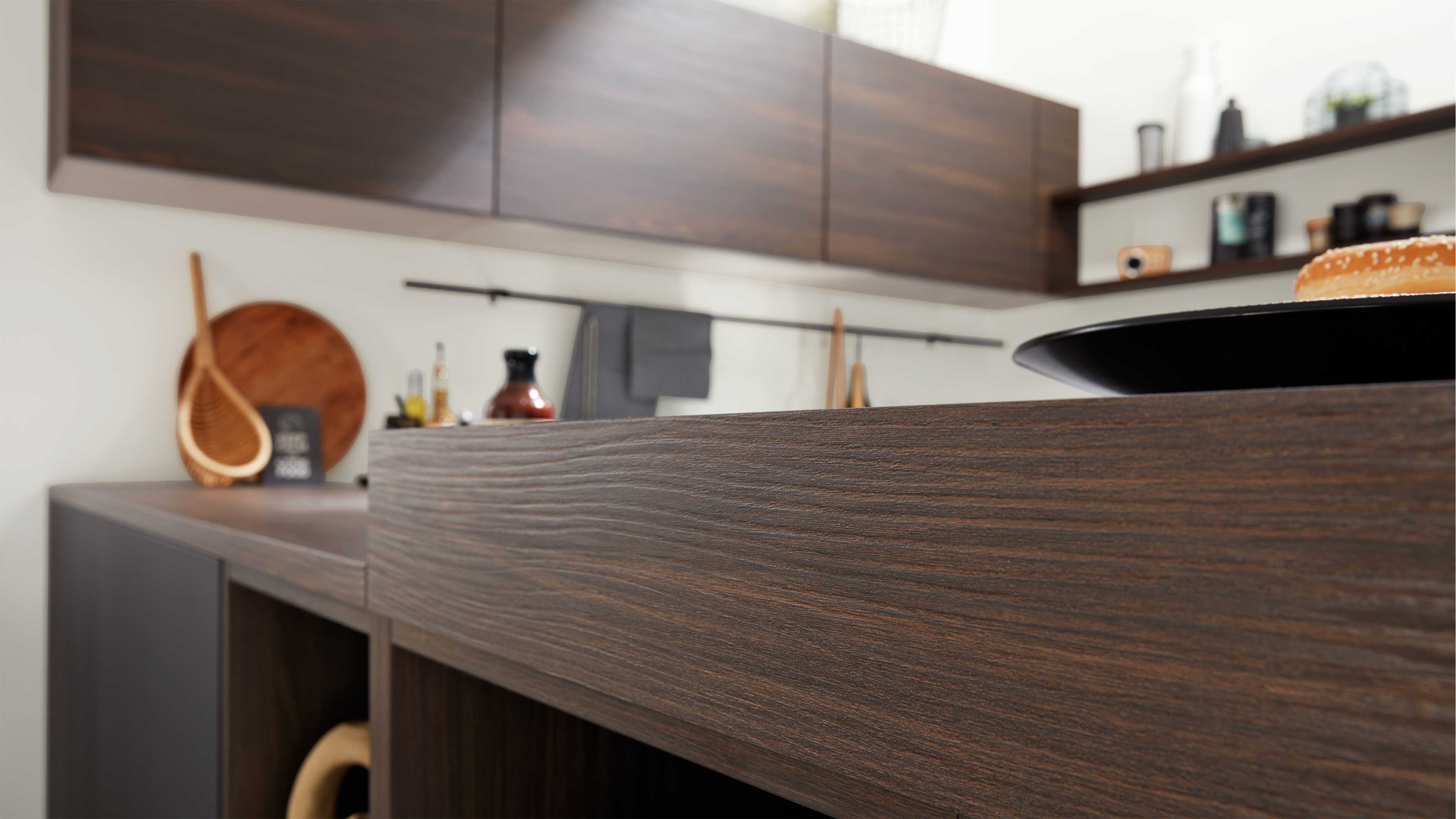 Dunkle Holzarbeitsplatte mit sichtbarer Maserung vorn, dahinter Oberschränke, Regale, Utensilien und ein Teller mit Brötchen.