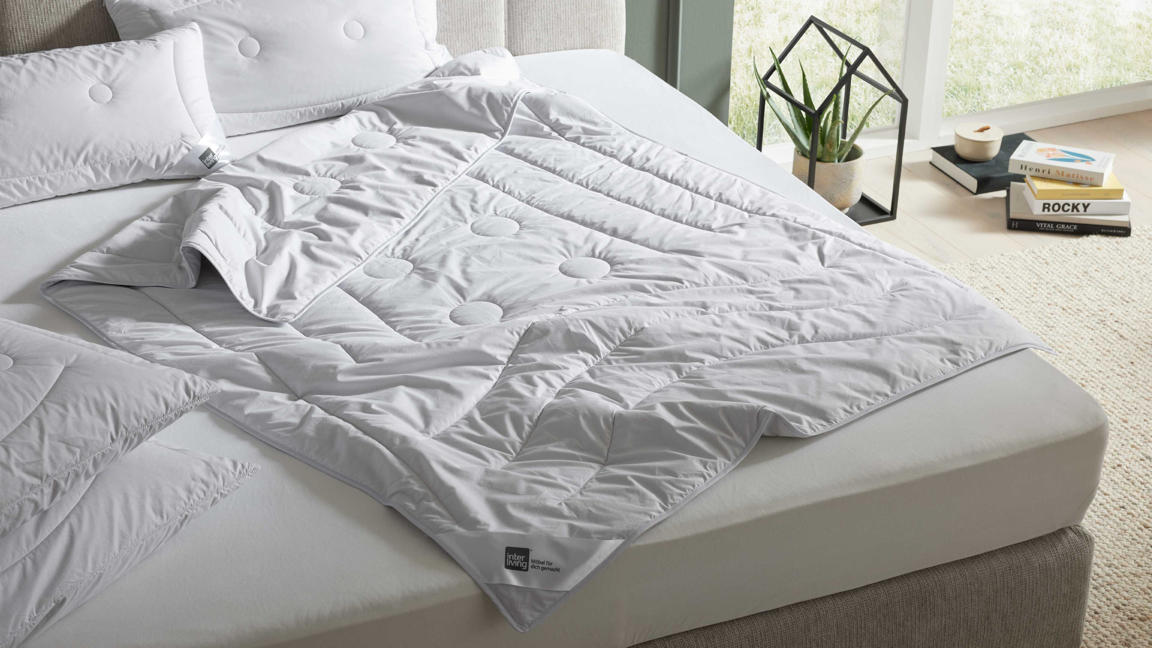 Weiße, gesteppte Bettdecke mit runden Stepppunkten liegt halb aufgeklappt auf hellgrauer Matratze, dazu zwei Kissen.