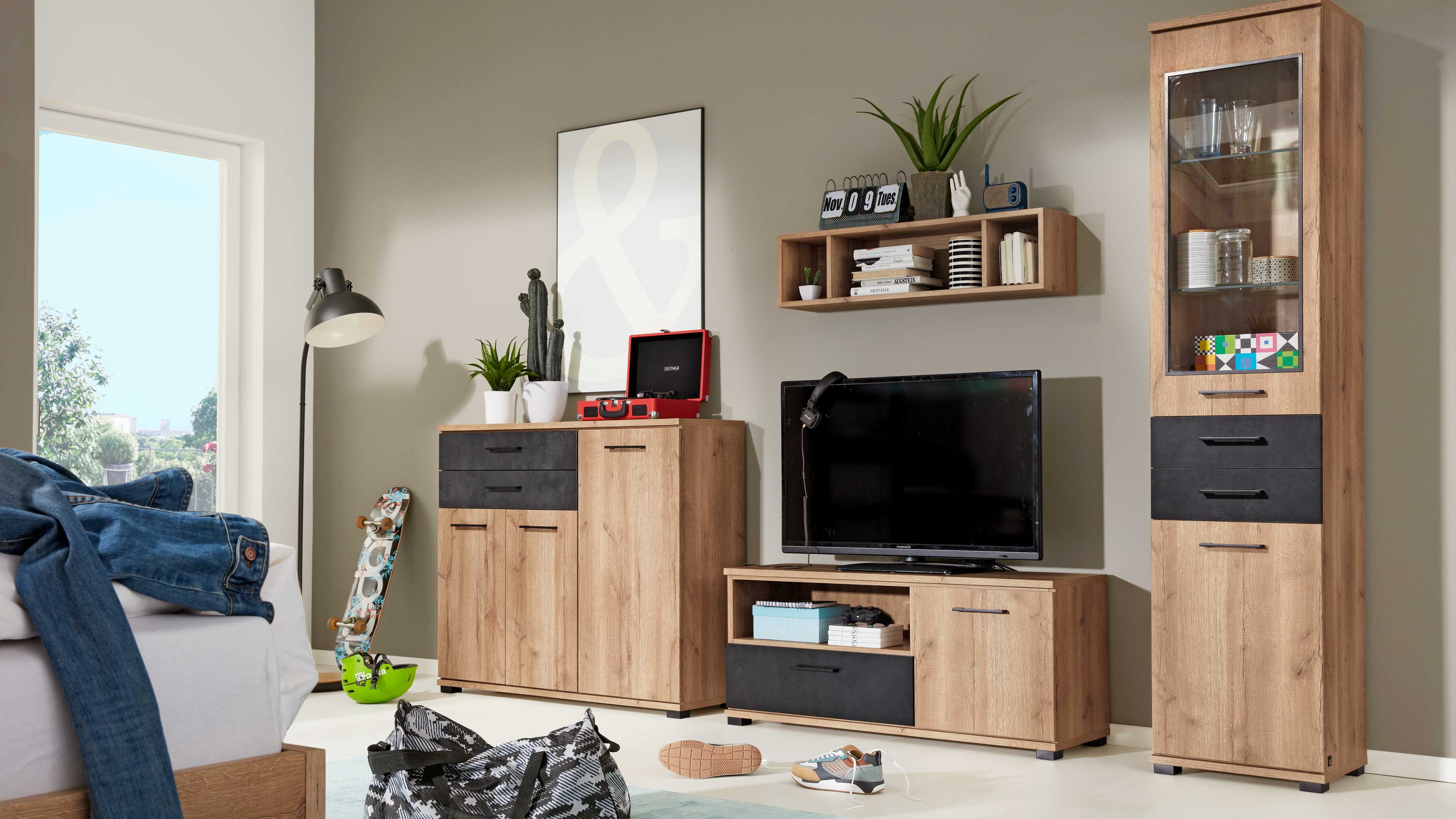 Wohnwand aus hellem Holz mit TV-Element, hohem Vitrinenschrank, Kommode und Wandregal, dazu Pflanzen, Skateboard und Fernseher.