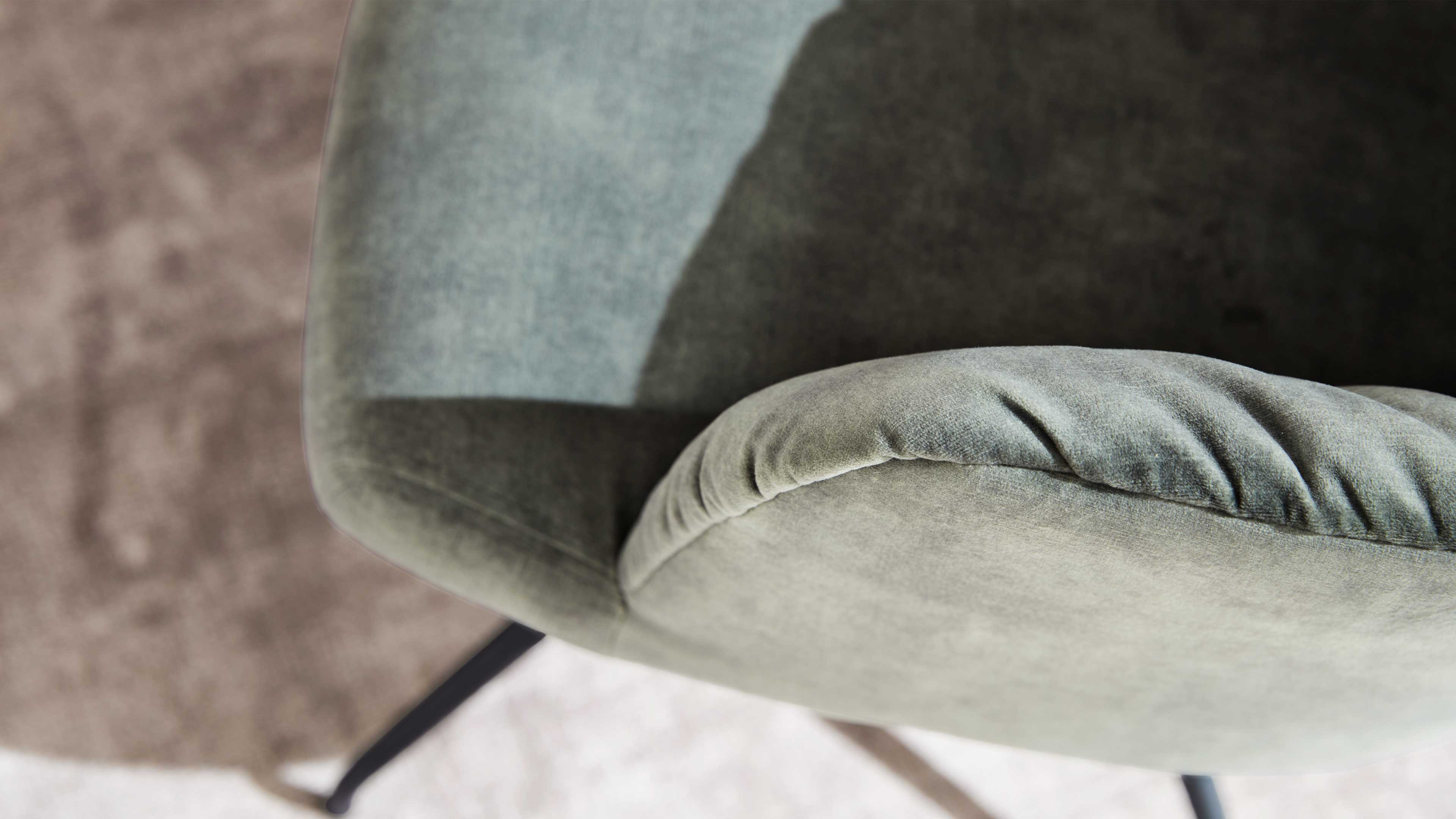 Gepolsterter, grünlich-grauer Sessel mit geschwungener Rücken- und Armlehnenkante, sichtbarem schwarzem Metallfuß und Teppich.