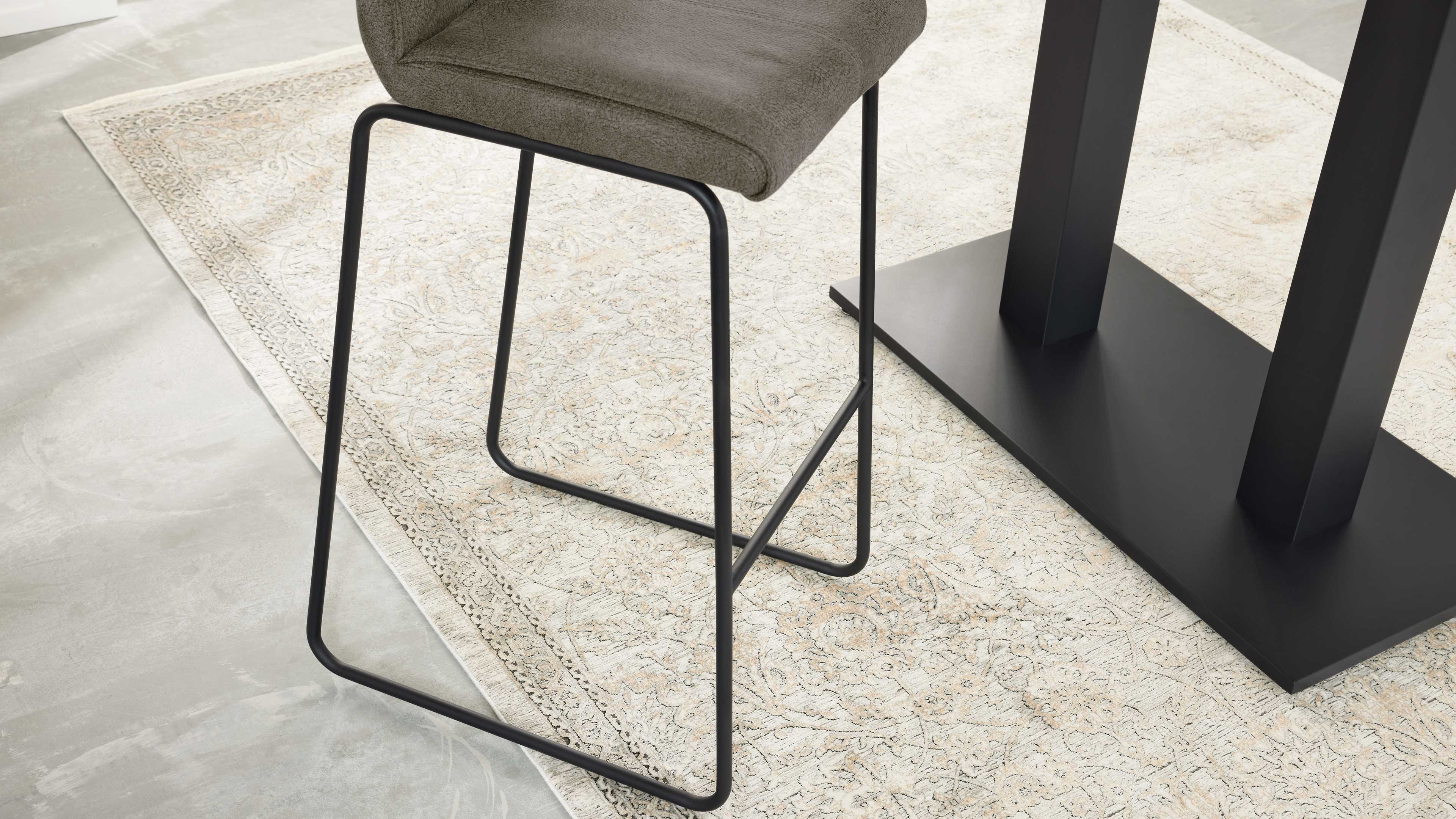 Hoher Hocker mit olivgrünem, gepolstertem Sitz und schwarzer Metallkufenstruktur auf beigem, ornamentalen Teppich neben schwarzem Tischfuß.