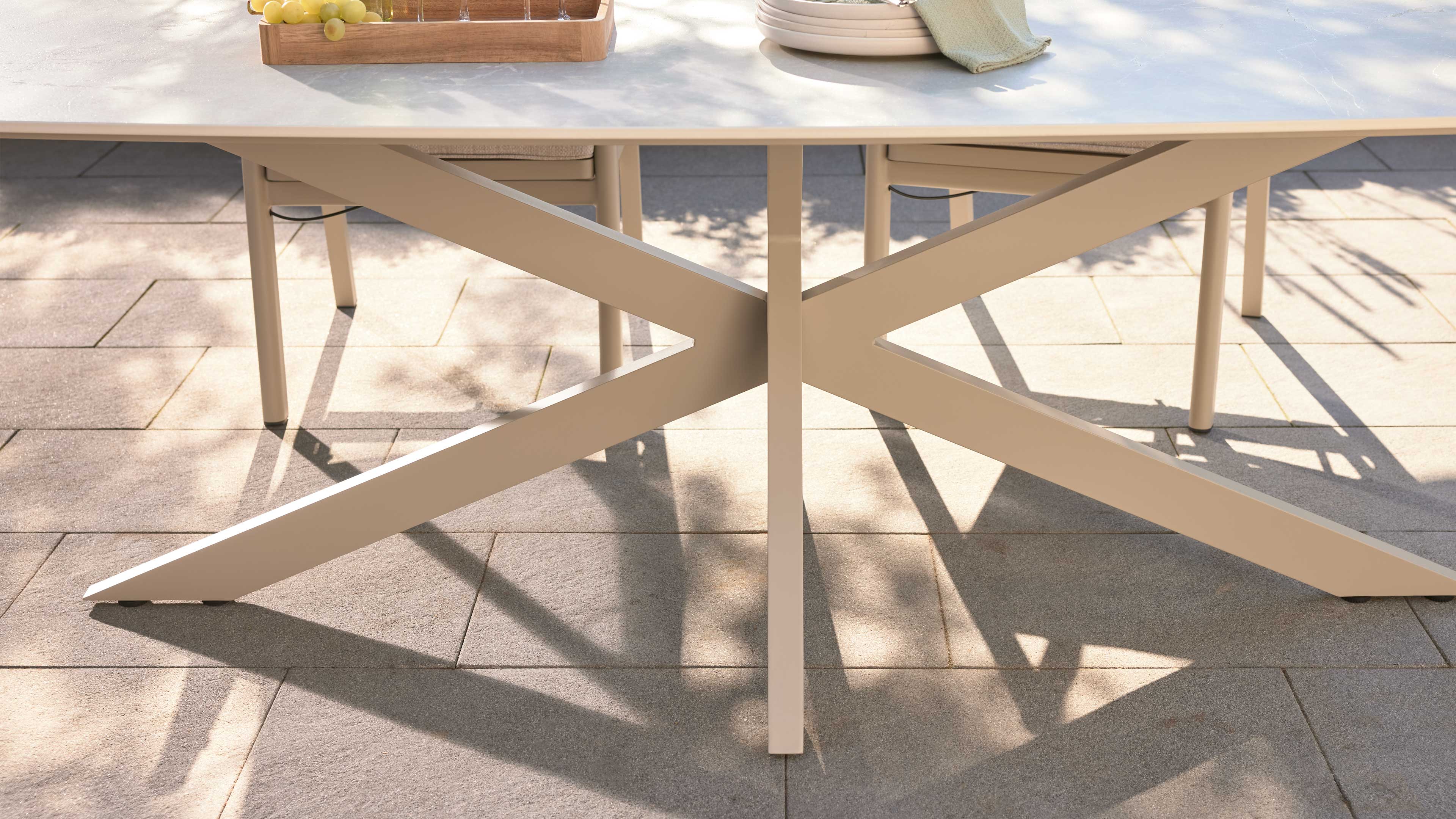 Beige Kreuzgestell eines Tisches mit einer rechteckigen Tischplatte, zwei Stuhlbeinen und Schatten auf hellen Terrassenplatten.