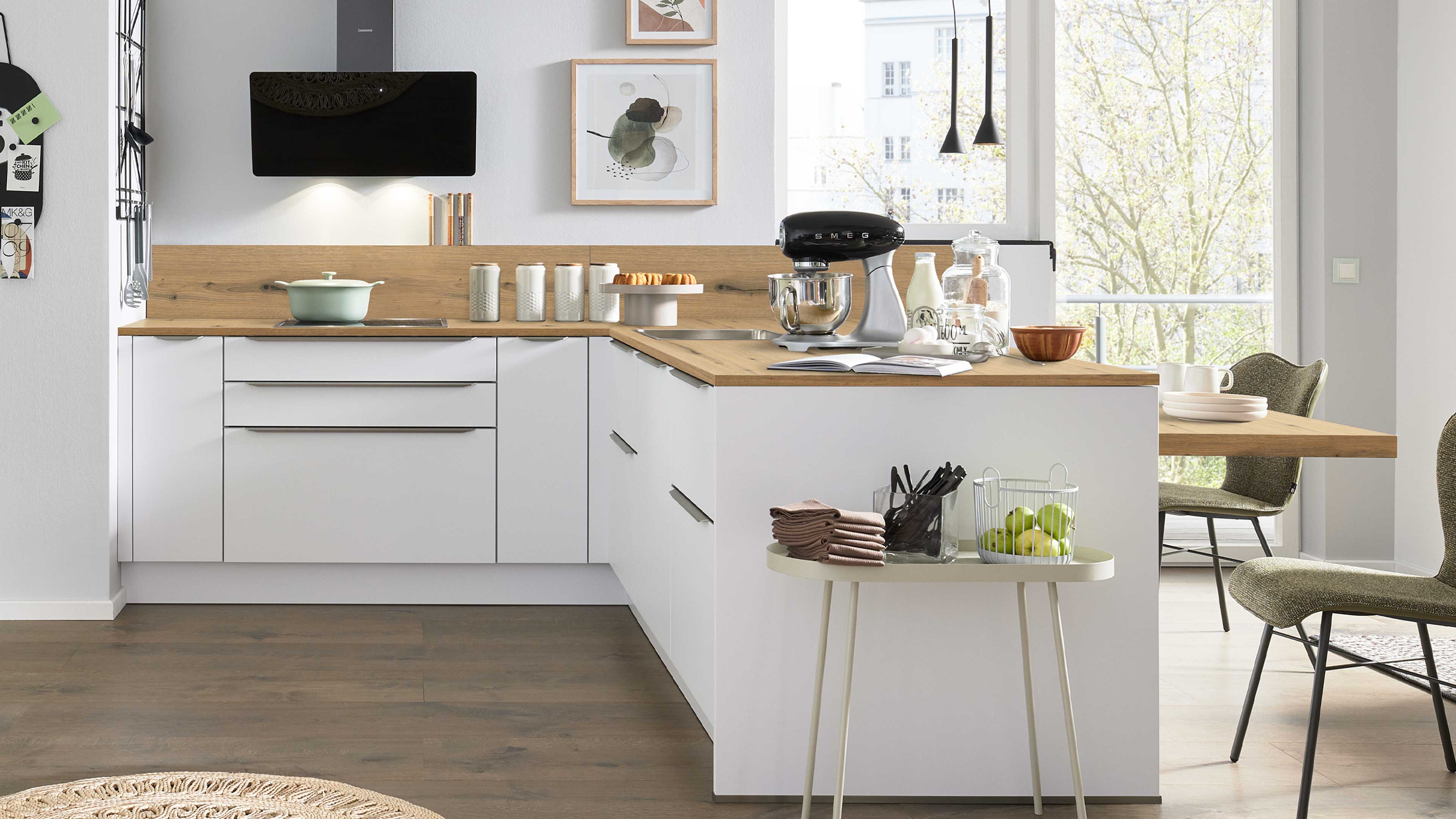Weiße L-förmige Küchenzeile mit Holzarbeitsplatte, schwarzem Standmixer, Gläsern, Obstkorb auf kleinem Beistelltisch und Stühlen.