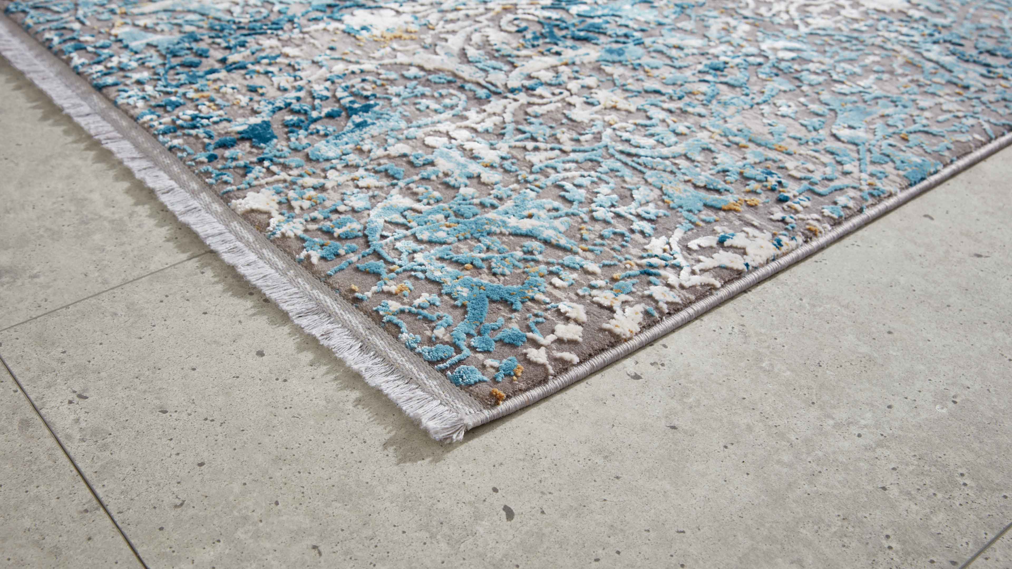 Eckansicht eines Teppichs mit grau-blau-weißem Reliefmuster, Bordüre und weißer Fransenkante auf hellgrauem Fliesenboden.