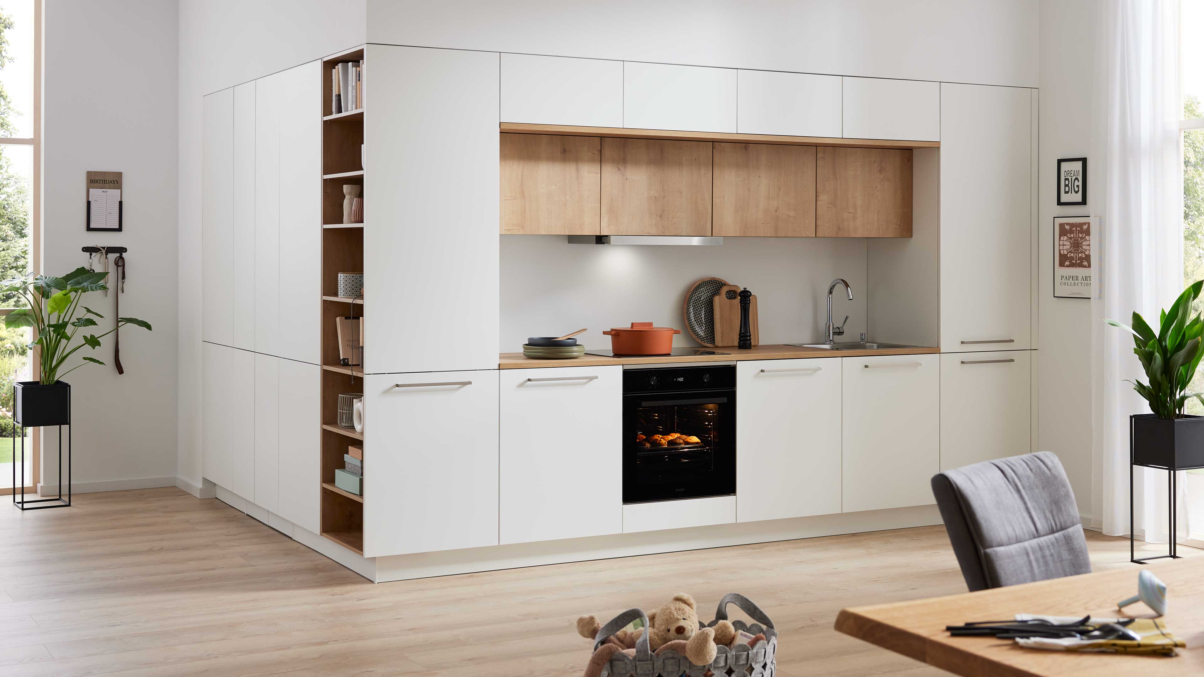 Weiße Einbauküche mit Holzarbeitsfläche und Oberschränken, Spüle, eingeschaltetem Backofen, offenem Regal und zwei Zimmerpflanzen.