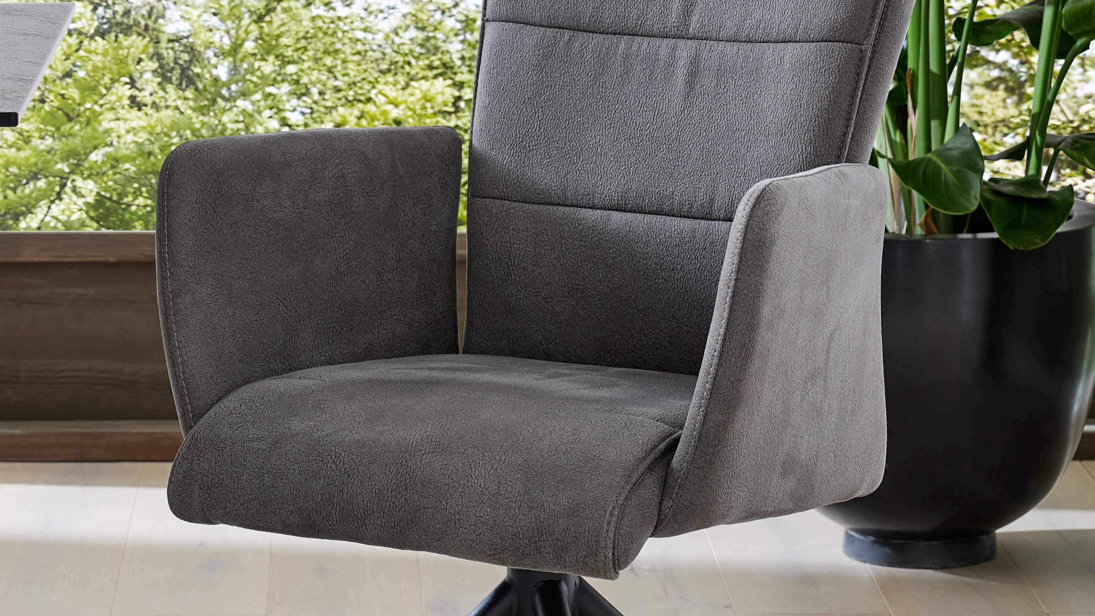 Grauer drehbarer Stuhl mit niedriger Rückenlehne und Armlehnen; rechts großer schwarzer Pflanzenkübel mit grünen Blättern, im Hintergrund Fenster.