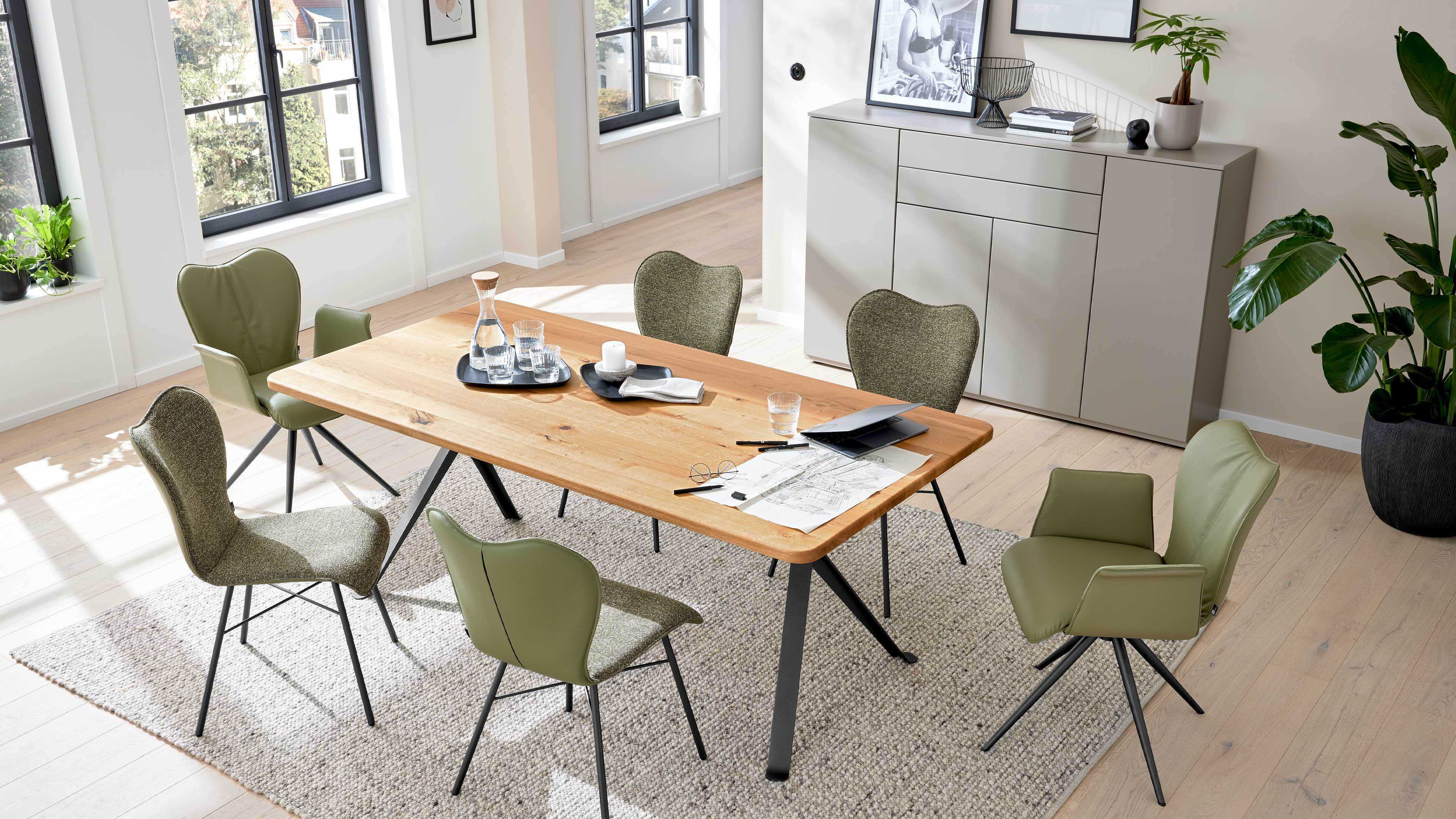 Esstisch aus hellem Holz mit schwarzen Metallbeinen, umgeben von sechs grünen Stühlen auf Teppich, Sideboard und Pflanzen.