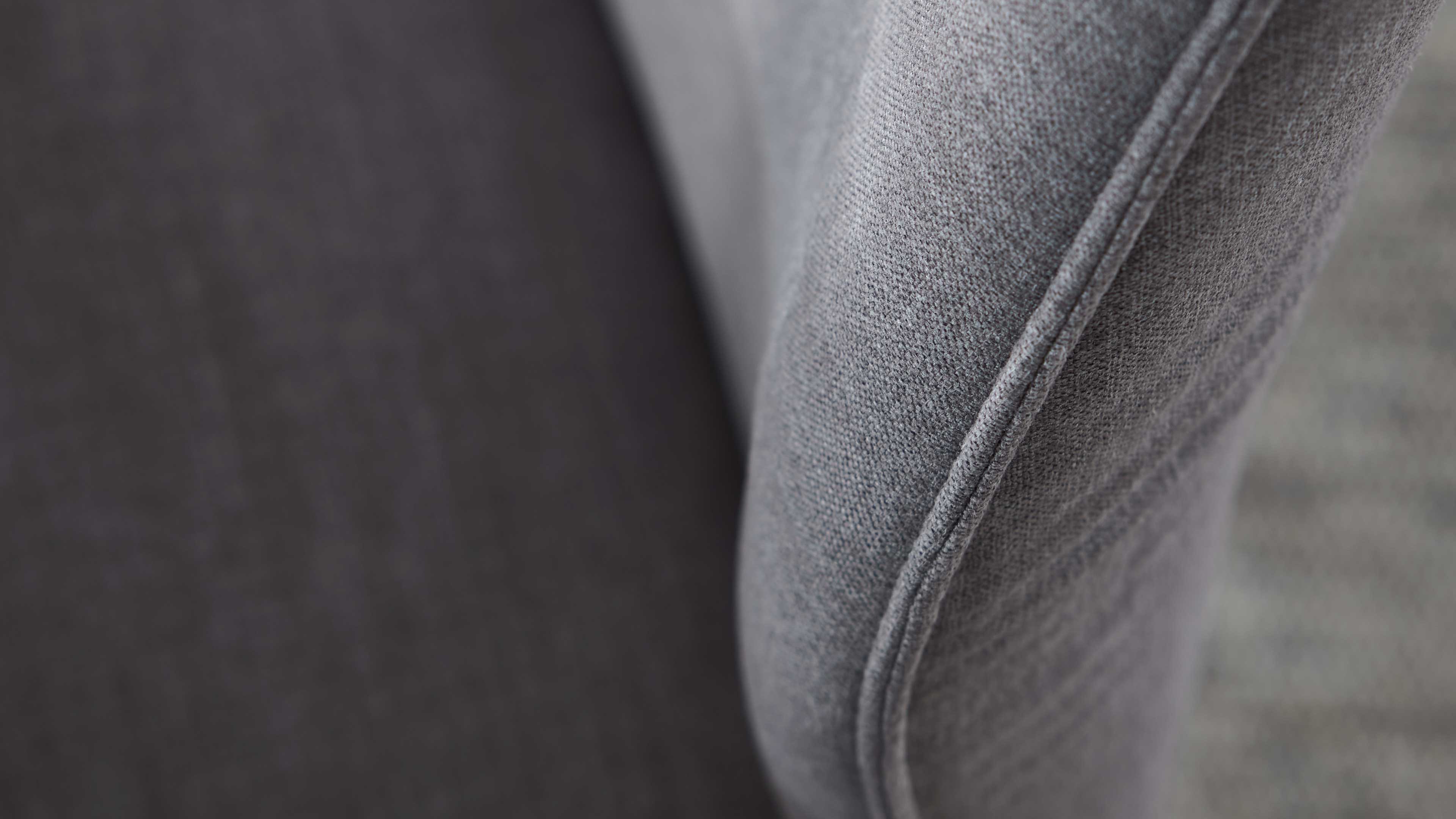 Nahaufnahme eines grauen Polsterbezugs mit sichtbarer Ziernaht am Armteil, daneben dunkelgraue Sitzfläche und gewebte Struktur.