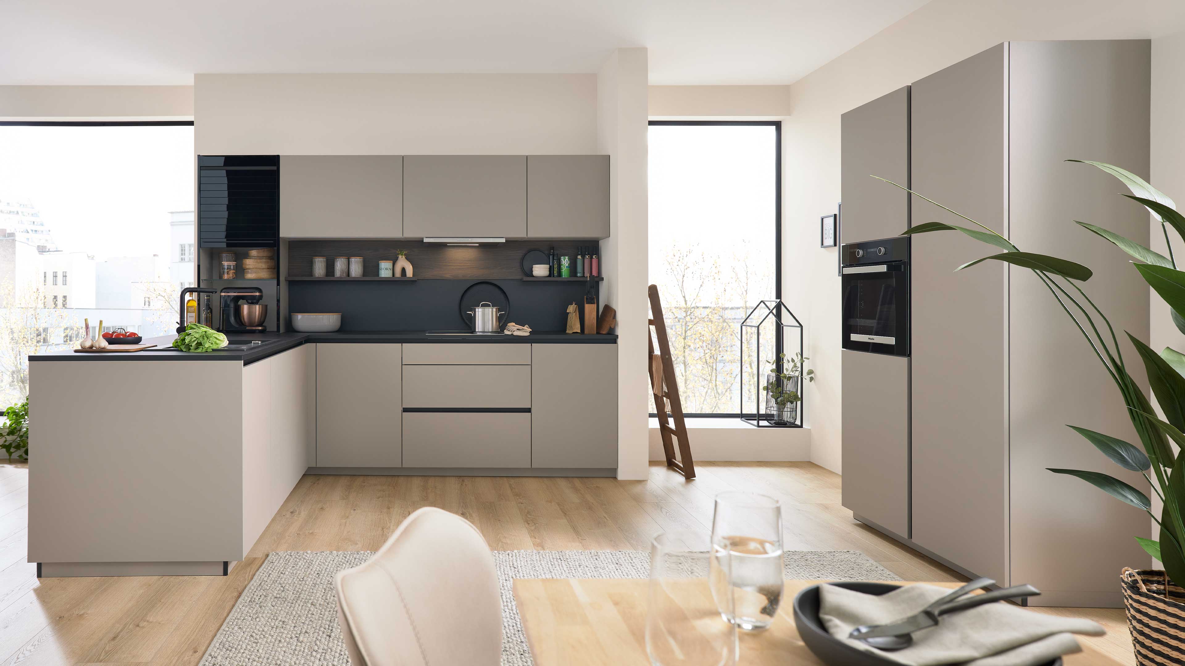 Beige L-förmige Küche mit schwarzer Arbeitsplatte und Insel, eingebautem Backofen, offenen Regalen, großen Fenstern und hellem Holzboden.