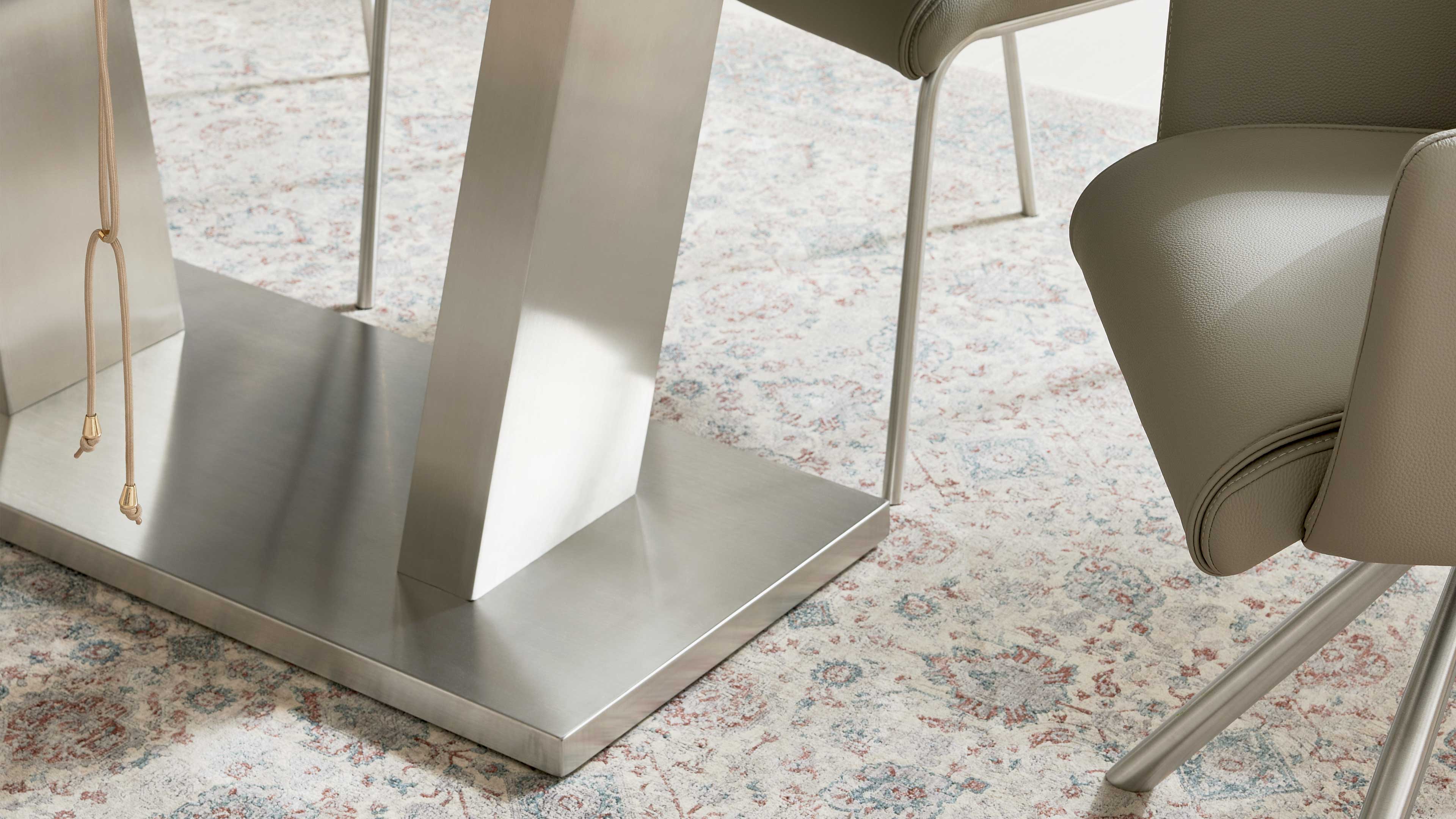 Breiter silberner Tischfuß auf rechteckiger Bodenplatte, daneben ein beigefarbener Polsterstuhl mit Metallbeinen und ein gemusterter Teppich.