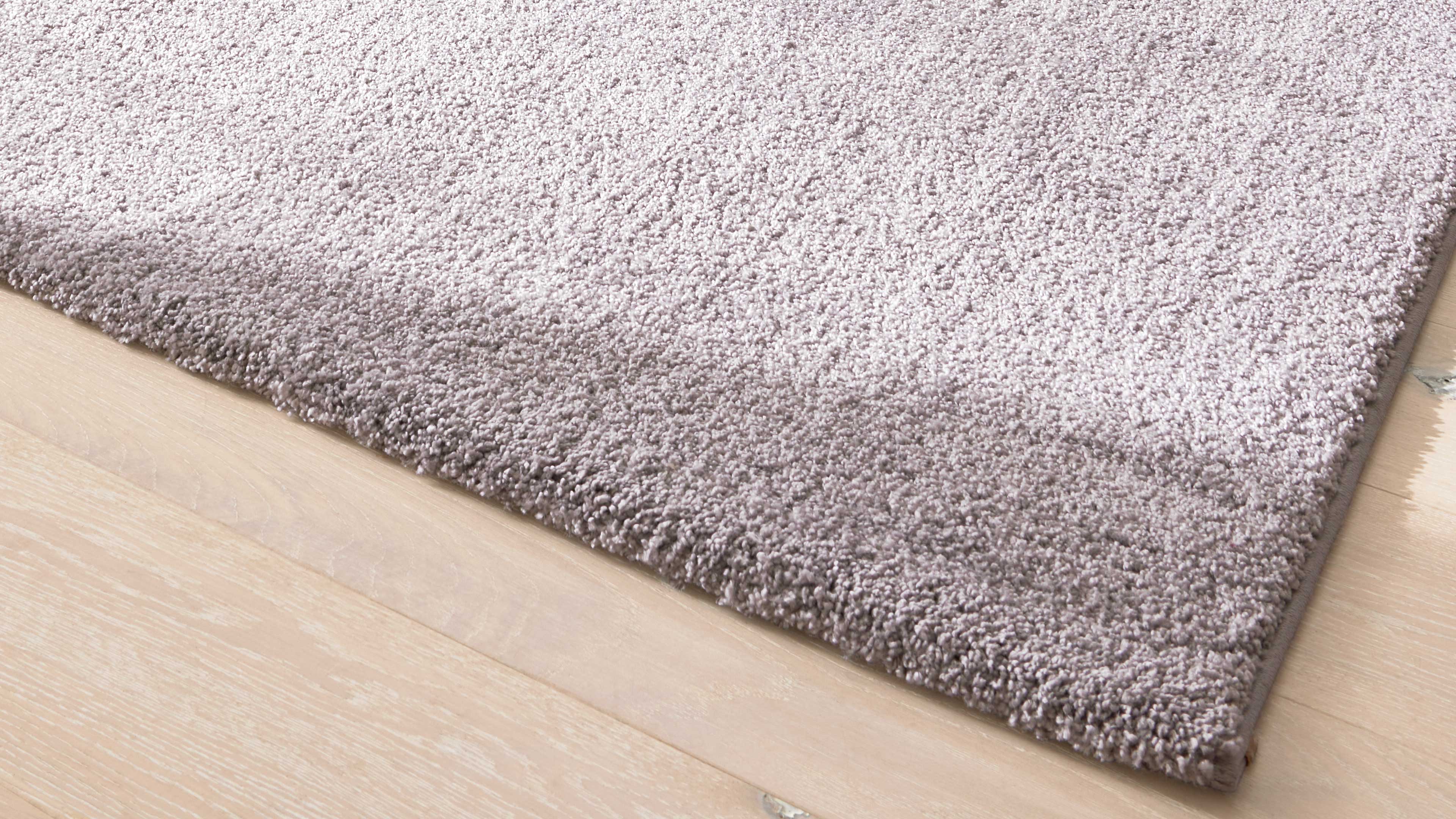 Hellgrauer, dichtfloriger Teppich liegt schräg auf hellem Holzfußboden, sichtbar eine abgerundete Ecke.