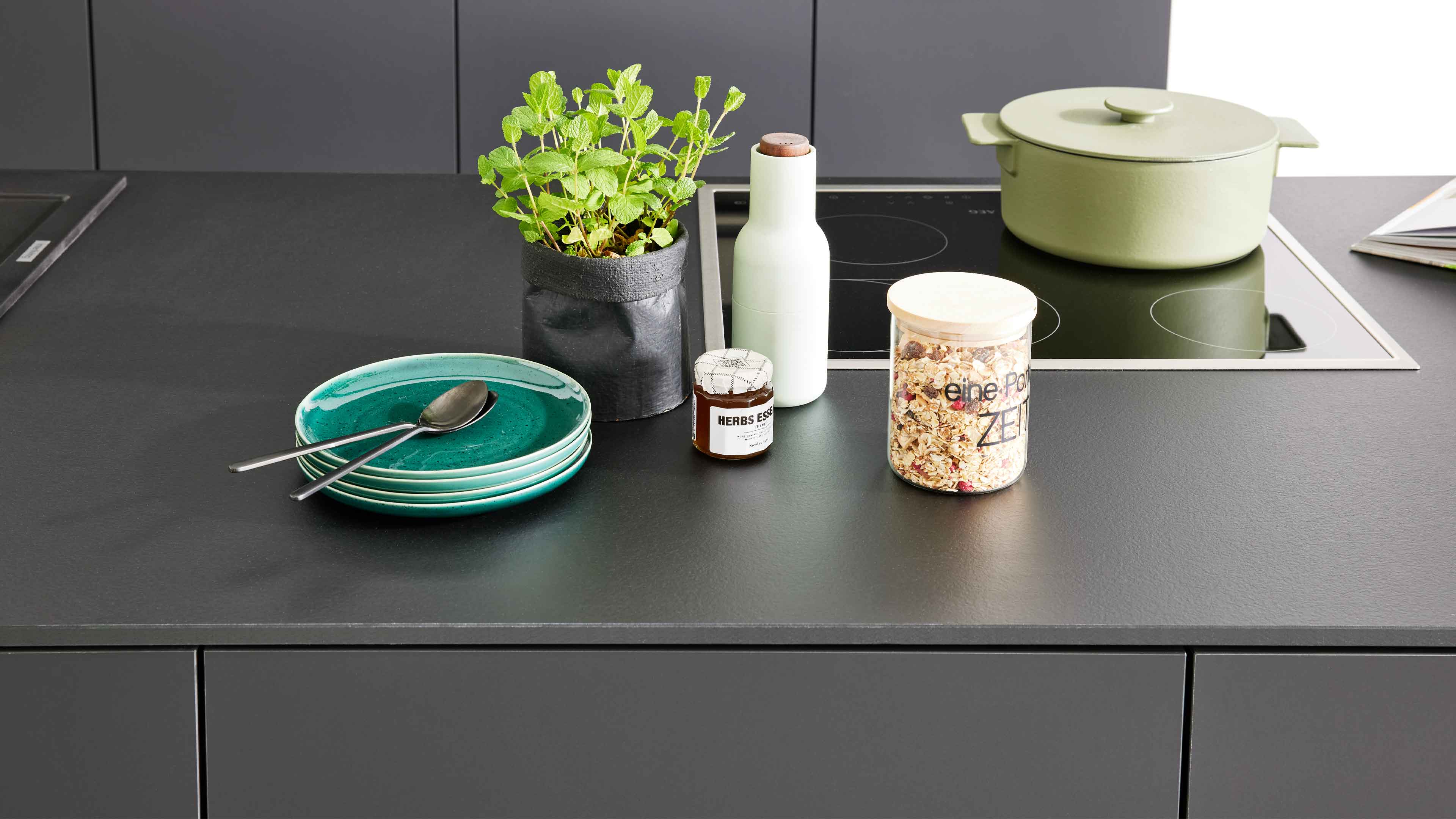 Dunkle Küchenarbeitsplatte mit integriertem Kochfeld, grünem Bräter, Müsliglas, gestapelten türkisen Tellern, Kräutertopf und Flasche.