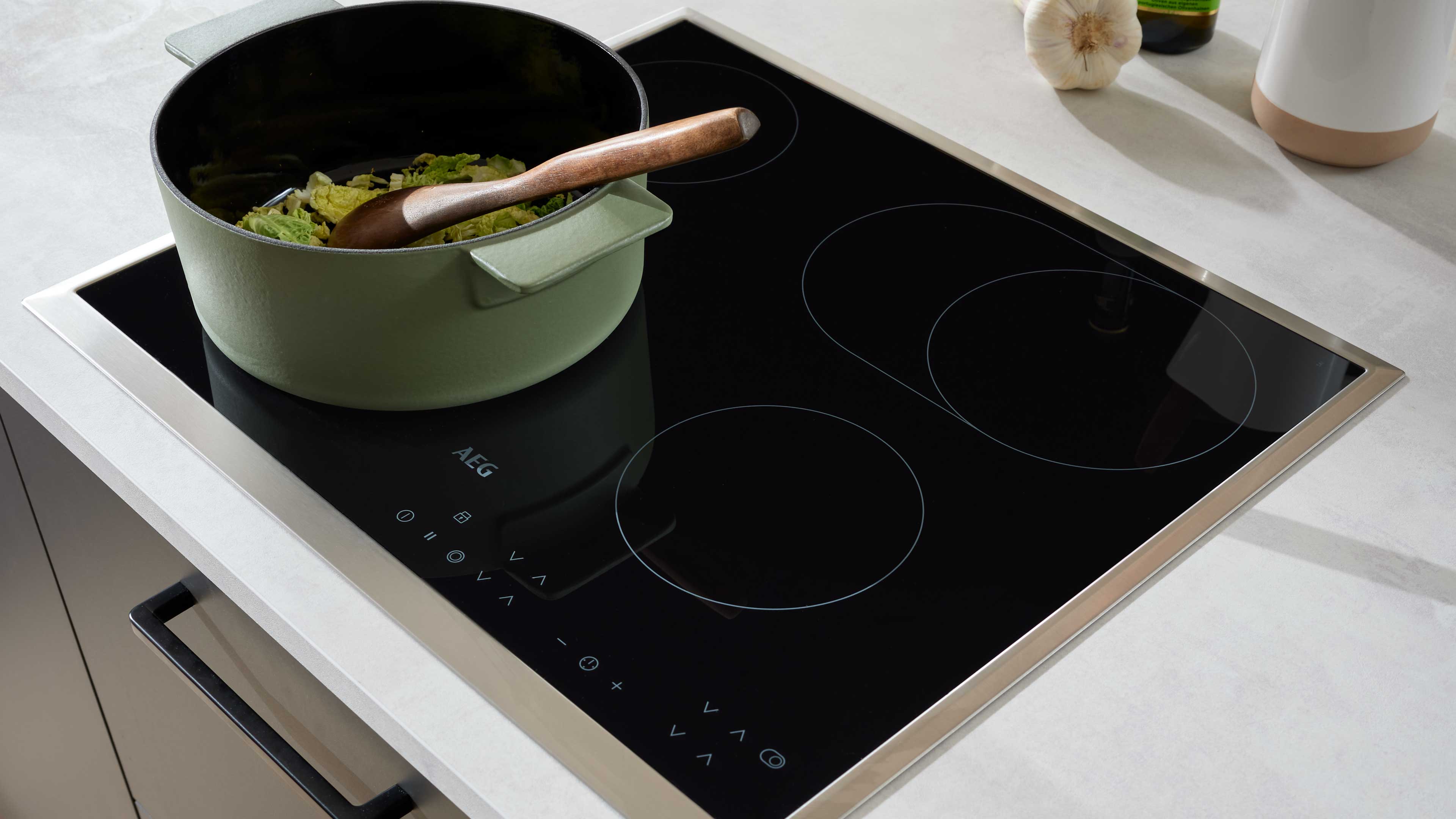 Schwarzes Glaskeramik-Kochfeld mit Edelstahlrahmen in heller Arbeitsplatte, darauf ein grüner Topf mit Holzlöffel und geschnittenem Kohl.