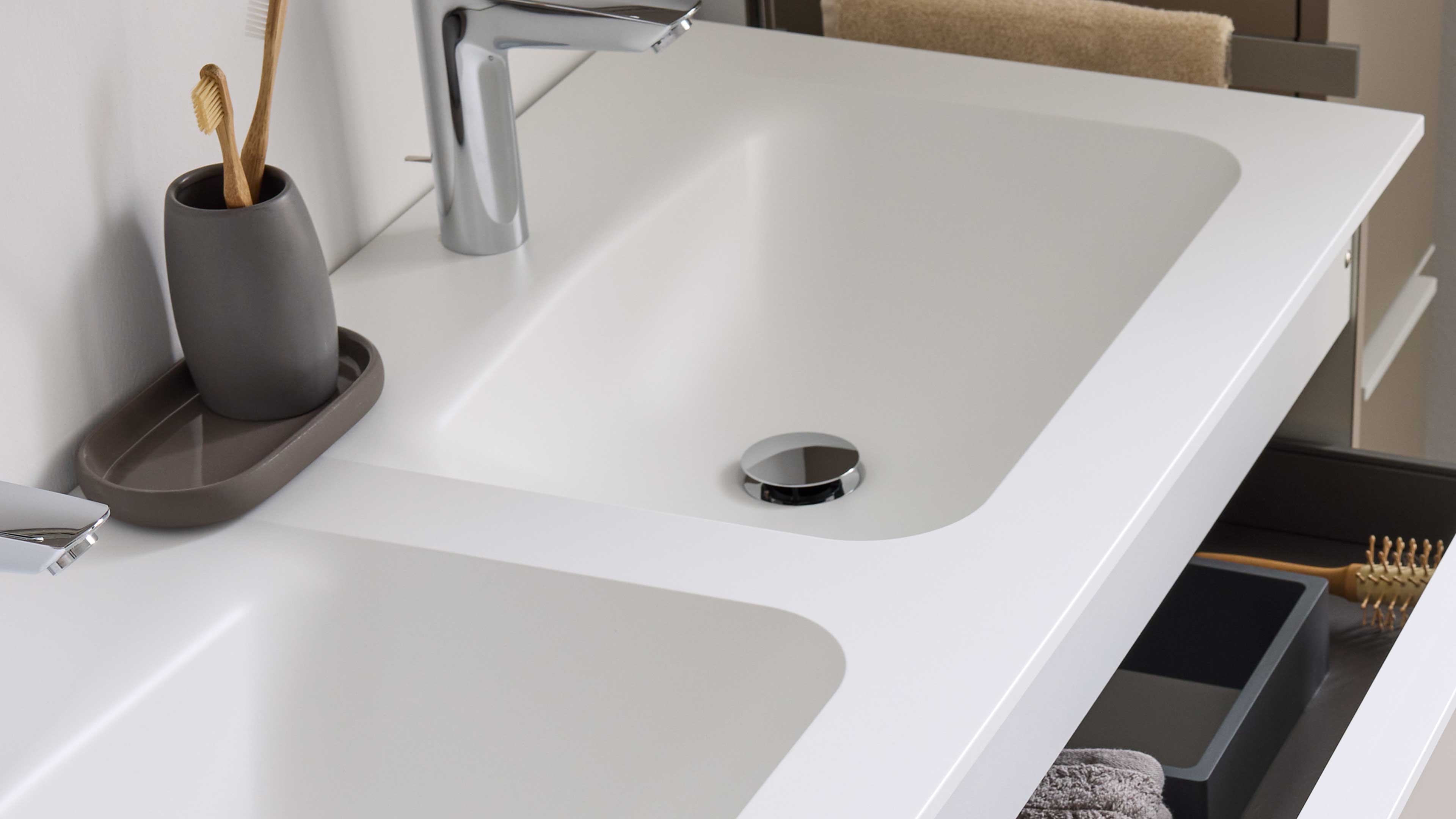 Weißer Doppelwaschtisch mit integrierten rechteckigen Becken, verchromten Armaturen, grauem Zahnputzbecher und offenem Schubfach.