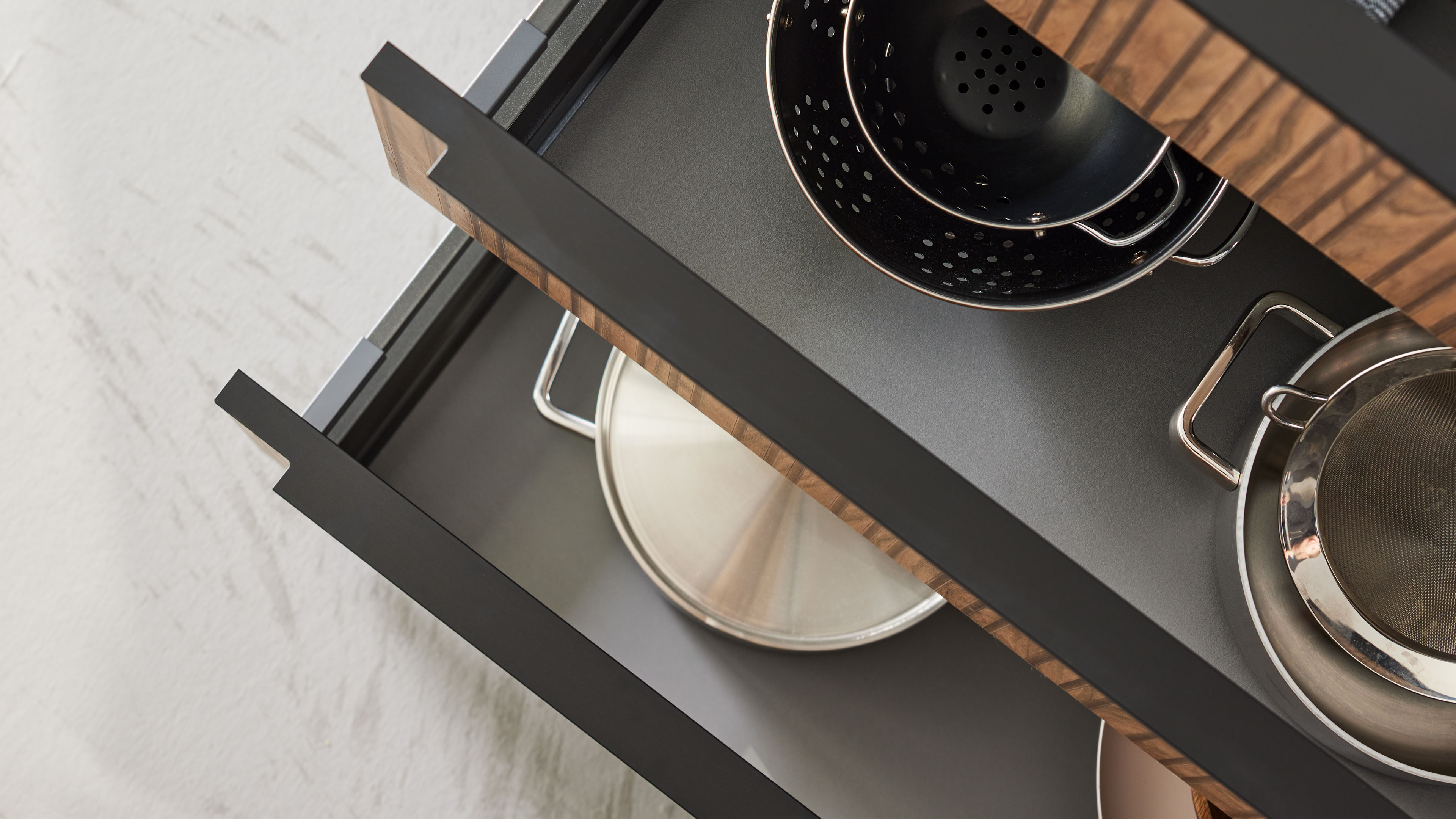 Offene Küchenschublade mit gestapelten Edelstahltöpfen, gelochtem Sieb und Seiher; schwarze Fronten mit Holzkante.