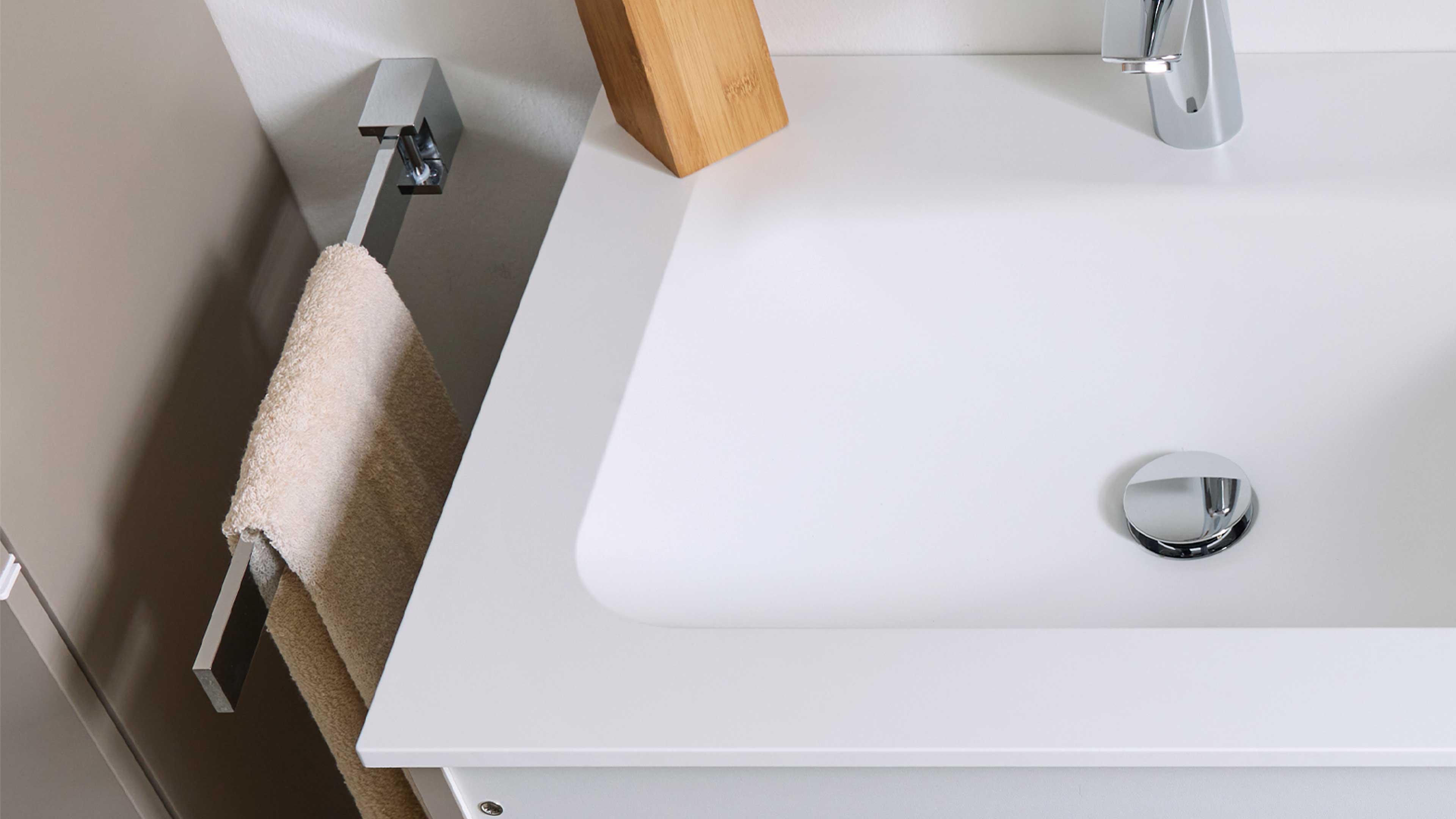 Weiße Waschbeckenplatte mit Becken, chromiertem Ablauf und Armatur oben rechts, oben mittig ein Holzblock, links ein beiges Handtuch.