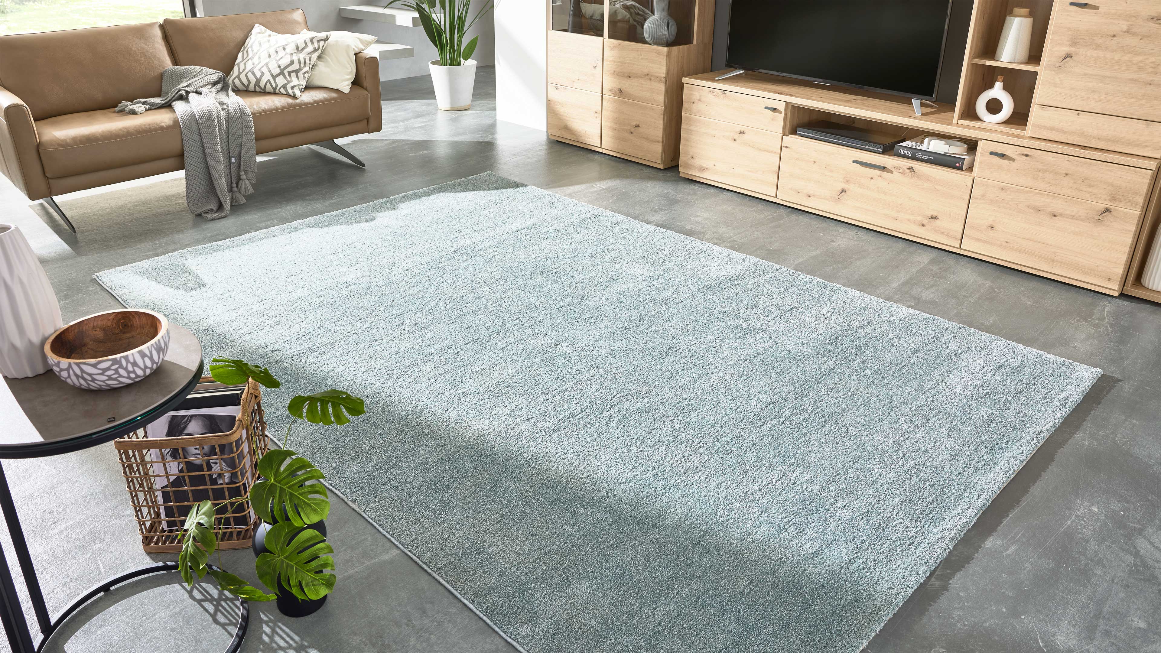 Großer hellblau-grauer Teppich auf grauem Betonboden, links cognacfarbenes Sofa, rechts helle Holzmedienmöbel mit Fernseher.
