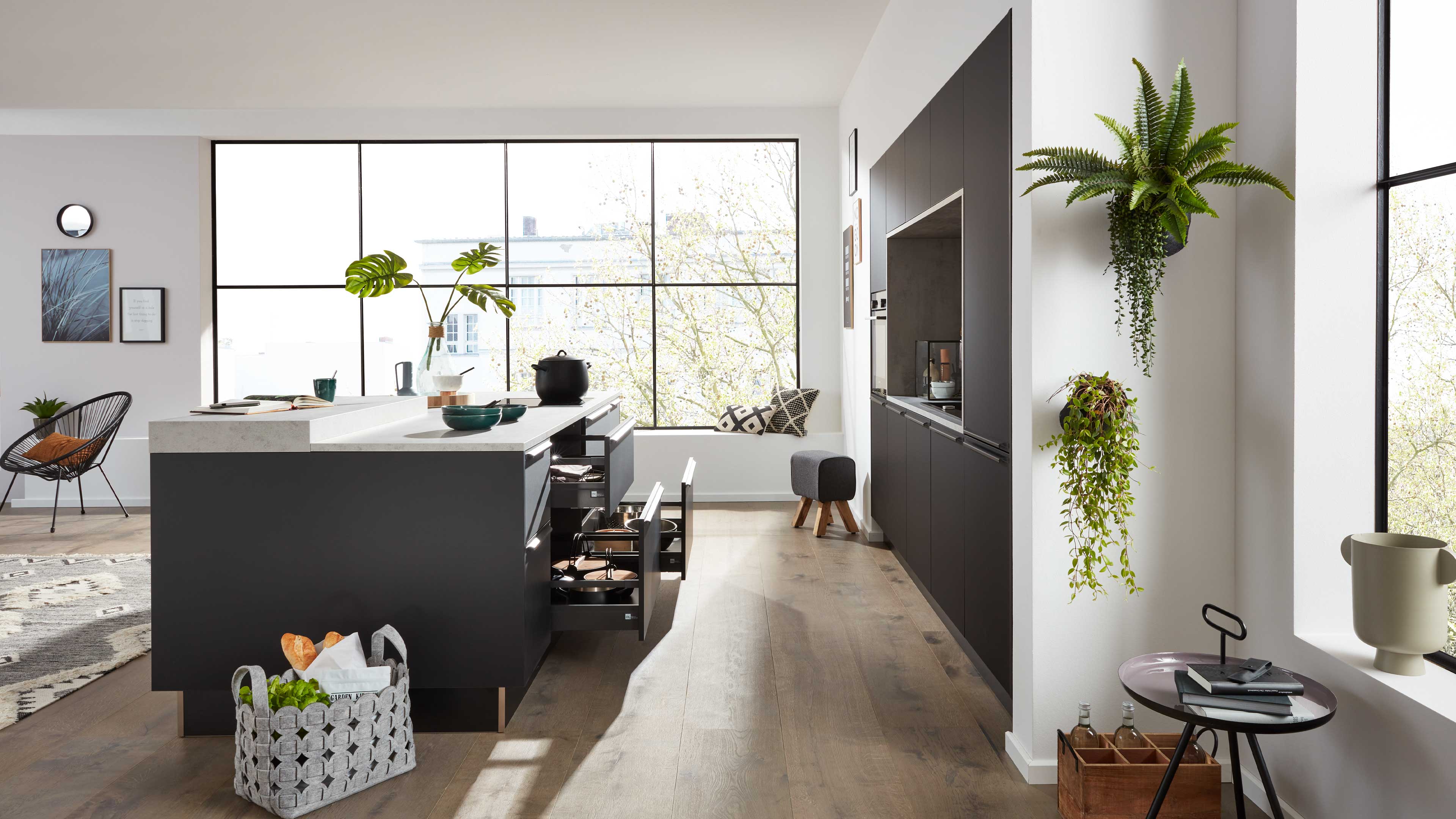 Dunkle Kücheninsel mit weißer Arbeitsplatte und geöffneten Schubladen, großes Rasterfenster im Hintergrund und hängende Pflanzen rechts.