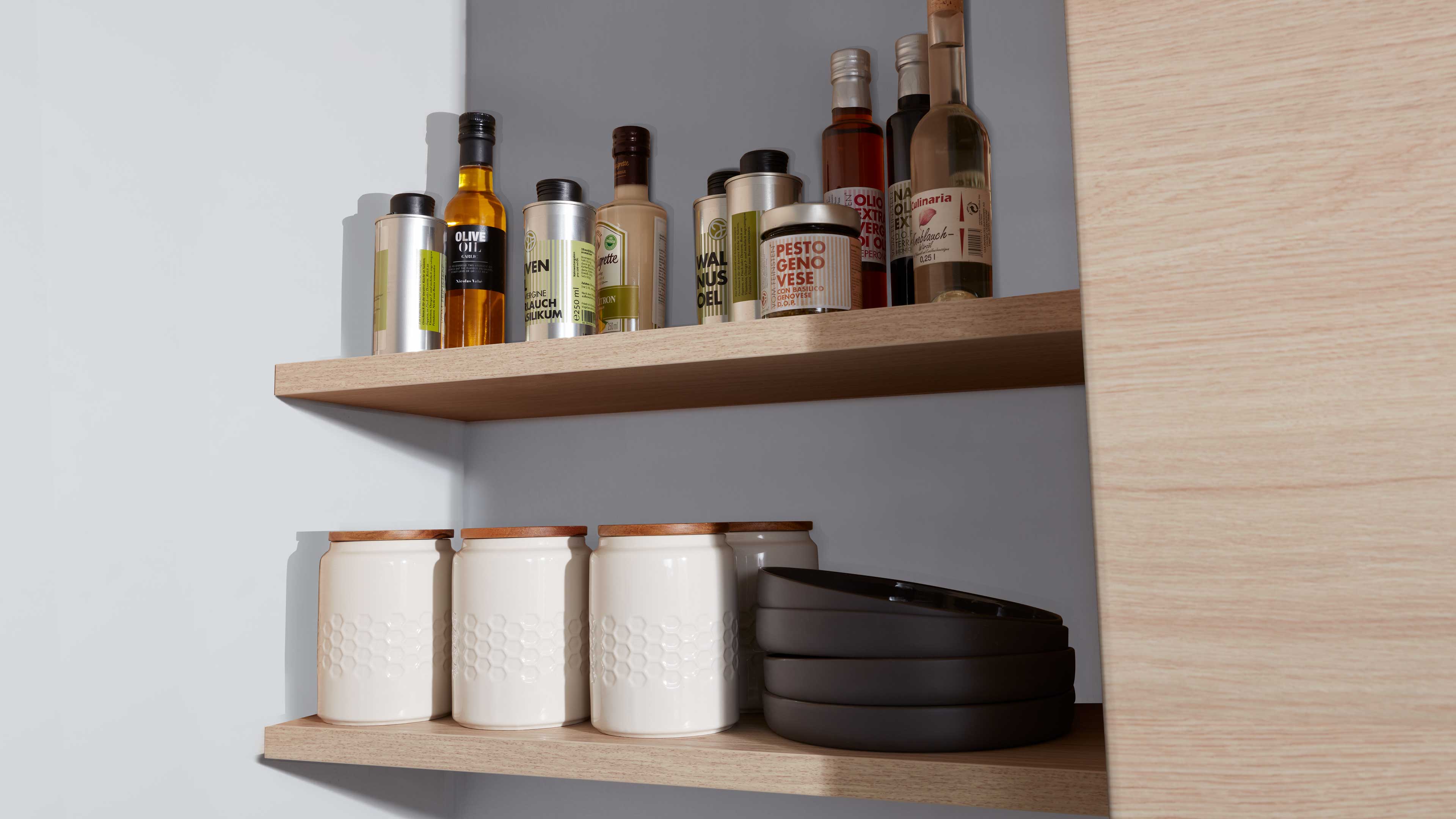 Helle Holzregalböden mit oben Öl- und Gewürzflaschen sowie Dosen, unten drei weiße Keramikdosen mit Holzdeckeln und dunklen Schalen.