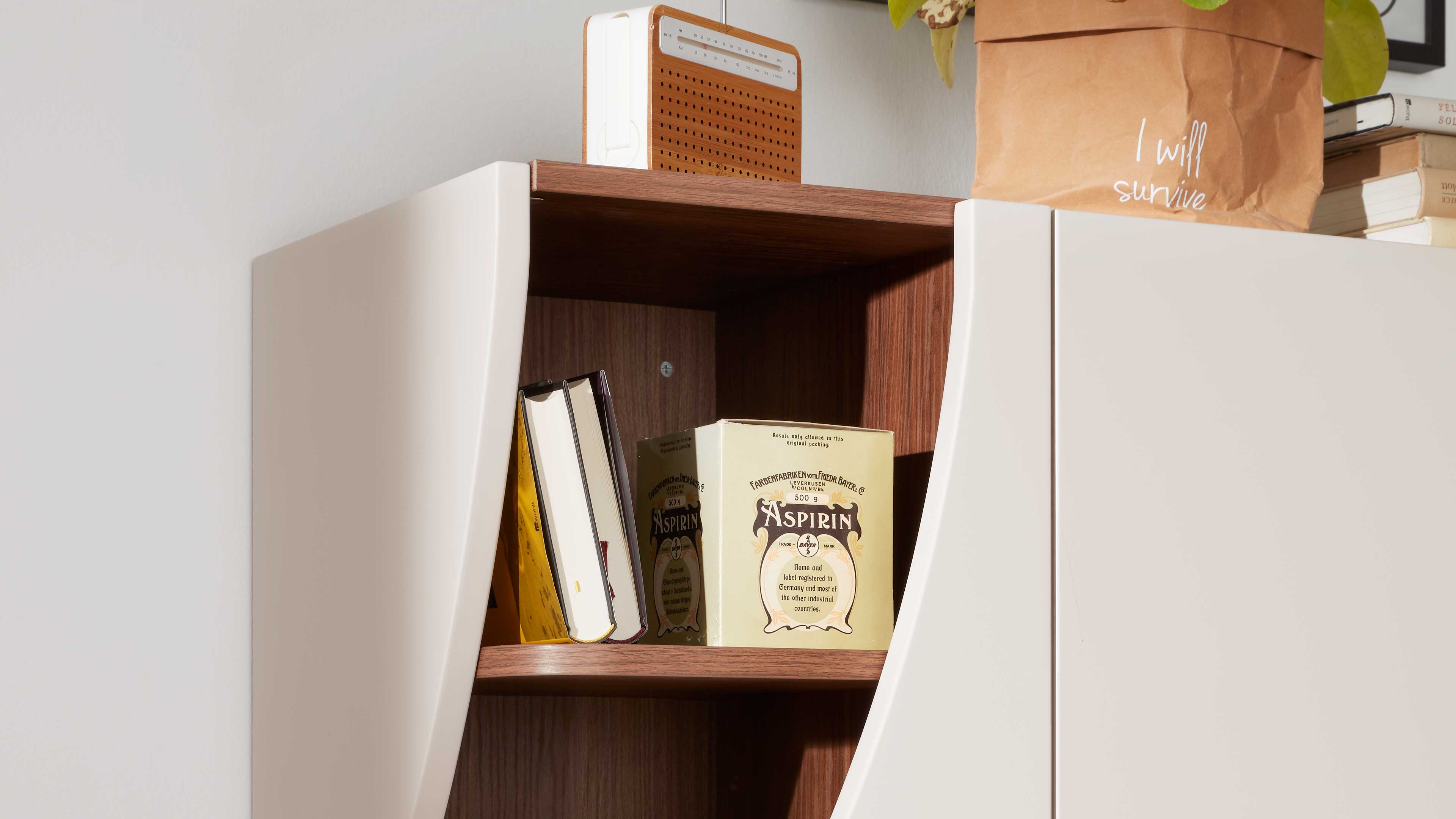 Wandhängender Schrank mit offener Mittelablage, weißen geschwungenen Türen und dunklem Holzinnenfach mit Büchern, Aspirin‑Dose und Radio.