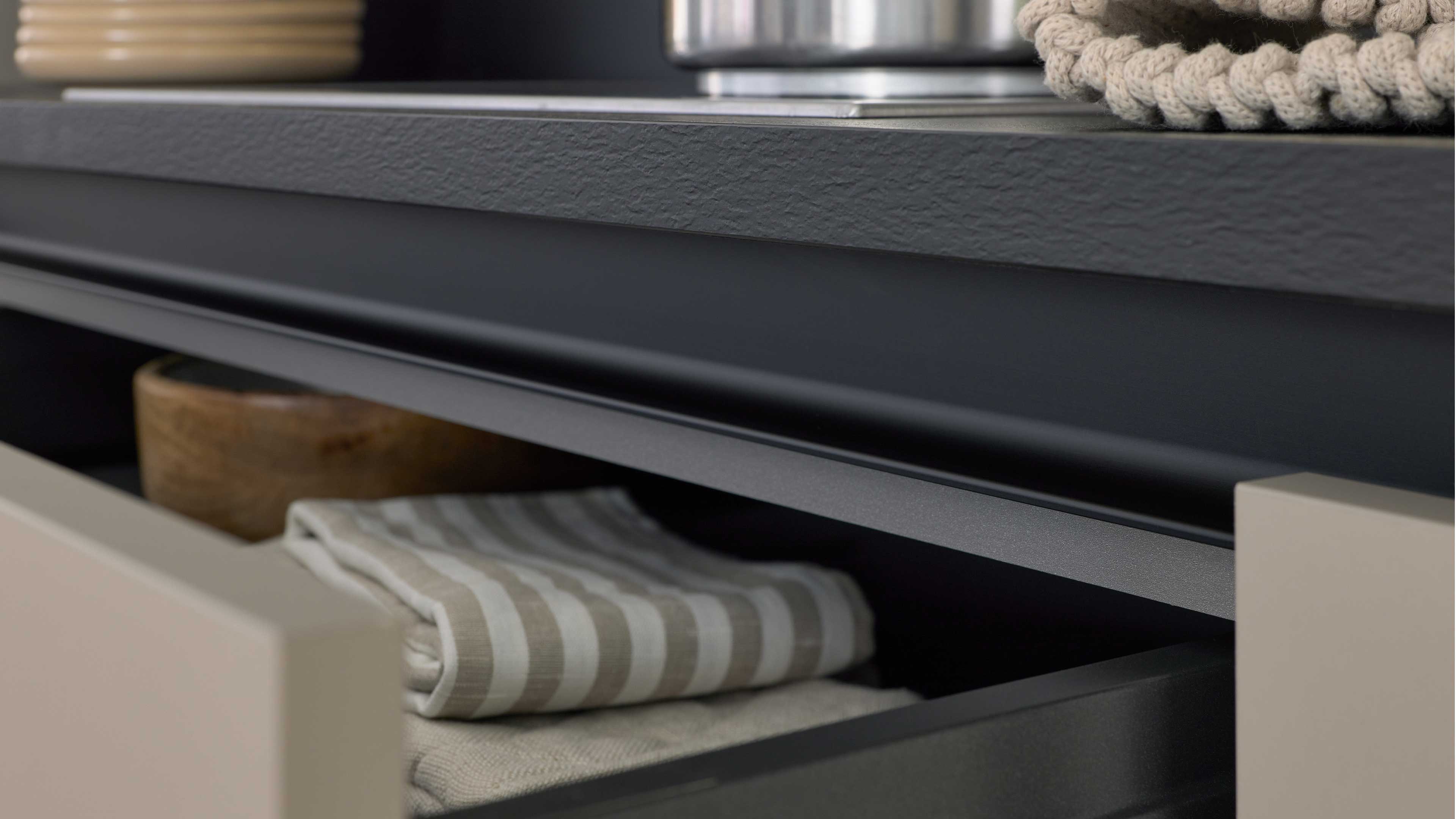 Offene schwarze Schublade unter einer strukturierten grauen Arbeitsplatte, innen gefaltete beige gestreifte Tischwäsche und eine Holzschale.