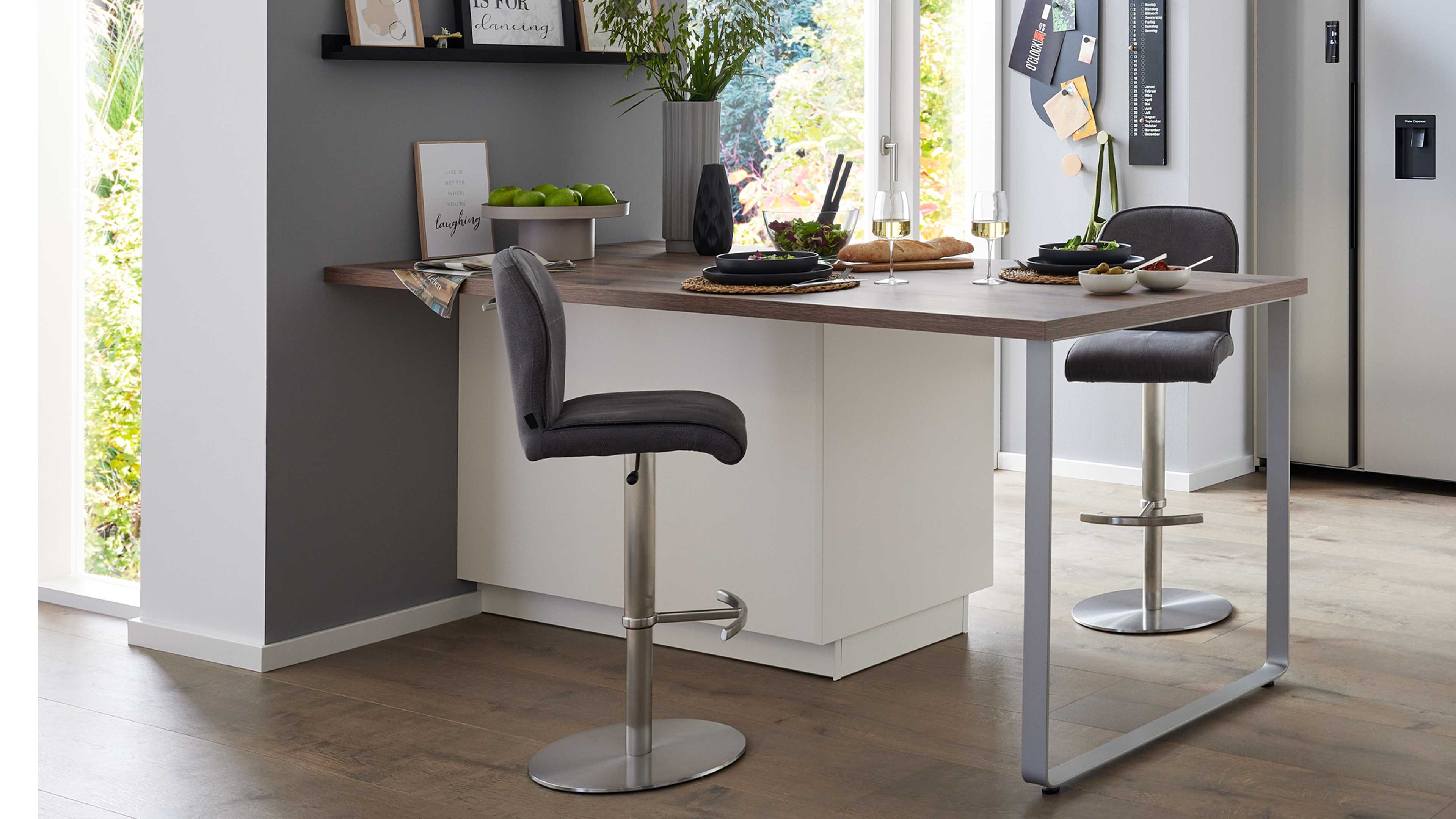 Barhohe Küchentheke mit braunem Holzplattenaufsatz auf grauem Metallgestell, davor zwei höhenverstellbare graue Polsterhocker.