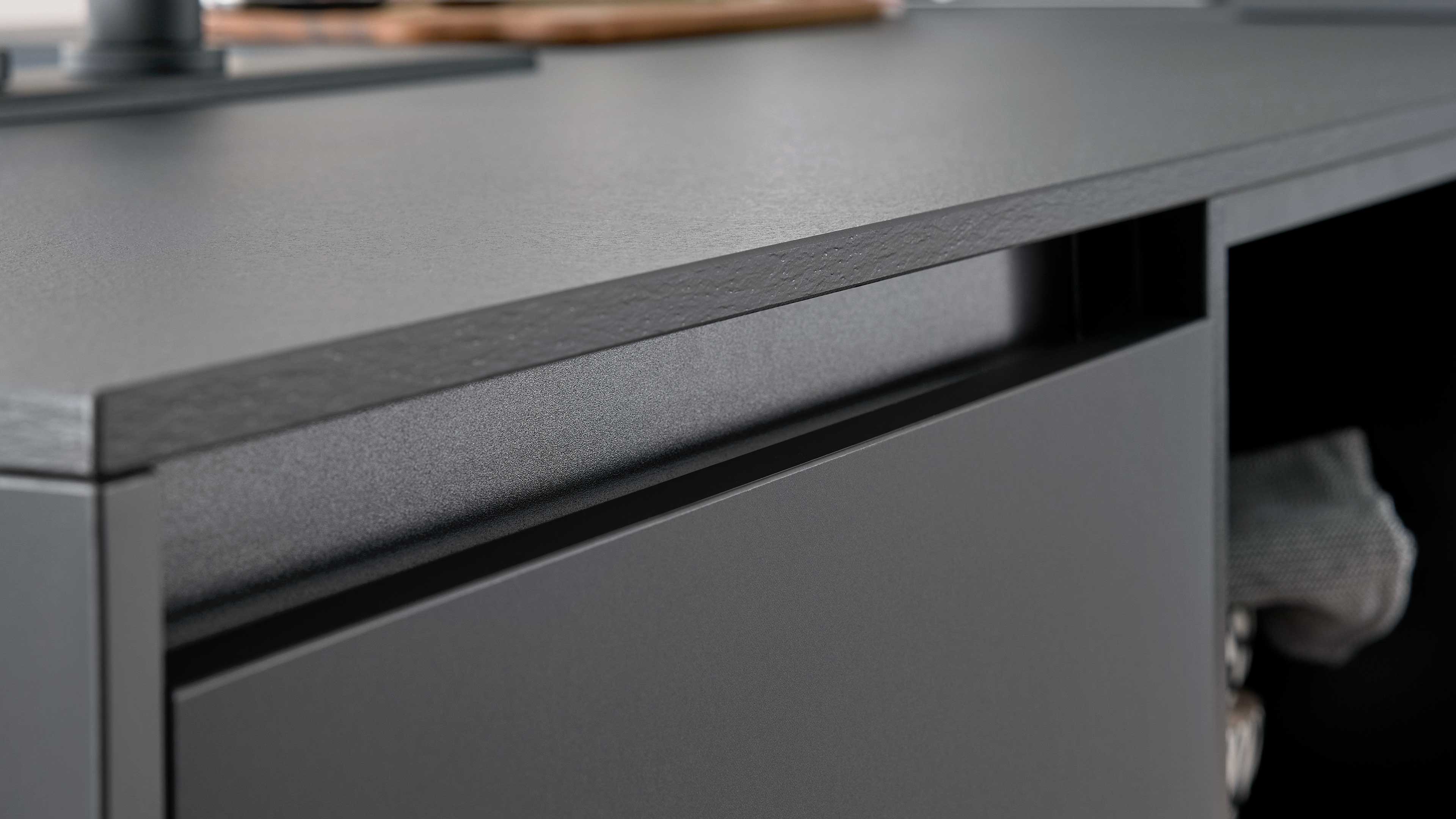 Dunkelgraue Schubladenfront mit eingelassenem Griff und strukturierter, matter Tischplattenkante, rechts offenes Fach mit Stofftuch.
