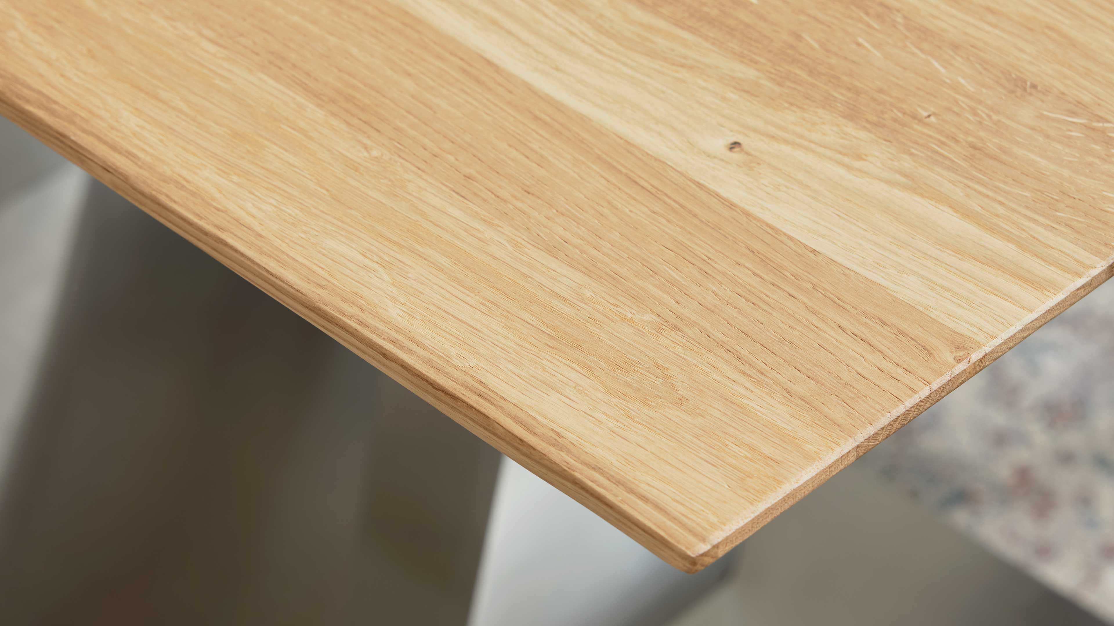 Eckansicht einer hellen Holz-Tischplatte mit sichtbarer Maserung, abgeschrägter Kante und teils sichtbarem weißen Tischbein.