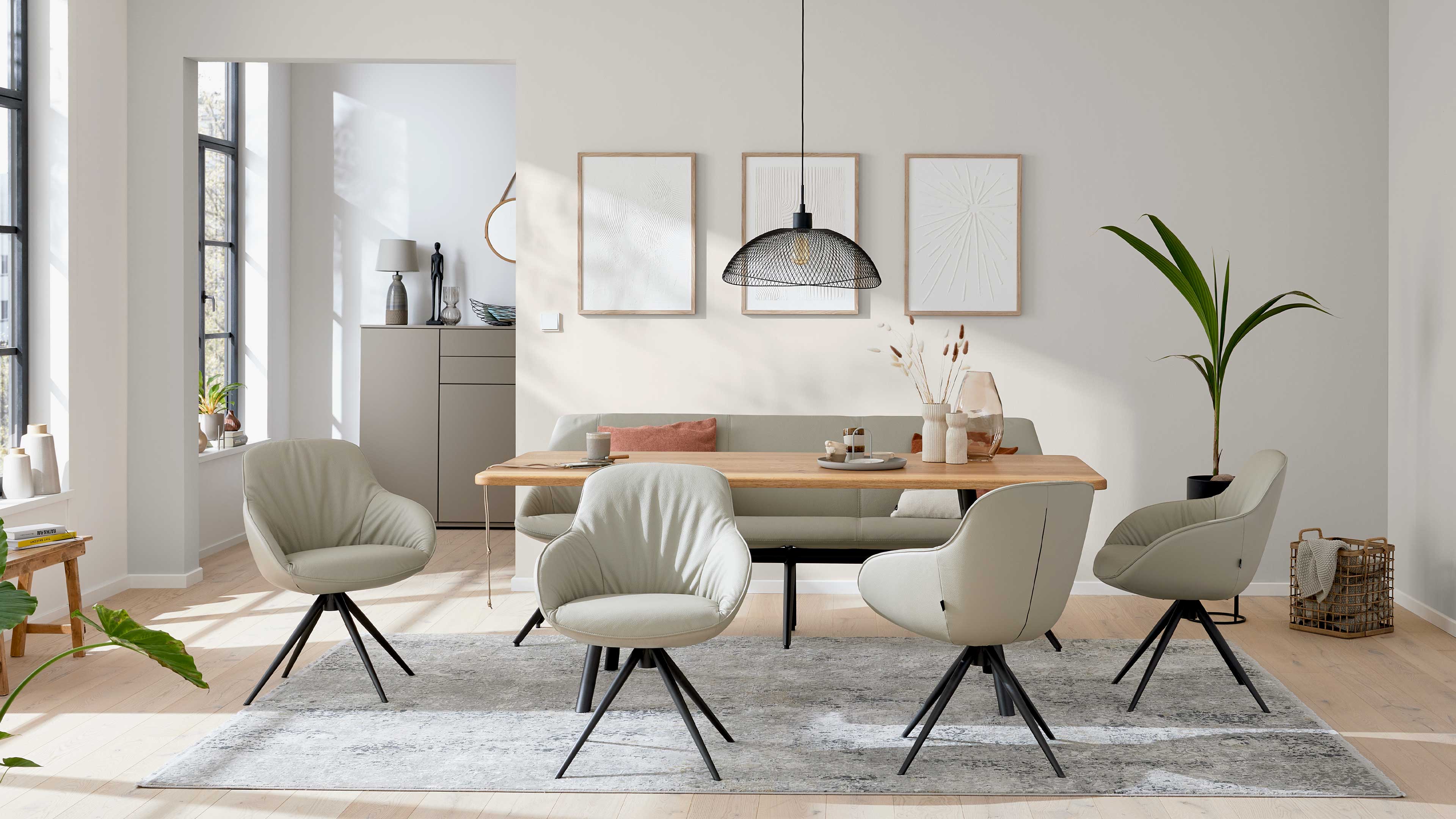 Esstisch aus hellem Holz mit vier beigen Drehstühlen auf grauem Teppich, Rückbank, Hängeleuchte, drei gerahmte Bilder und Topfpflanze.