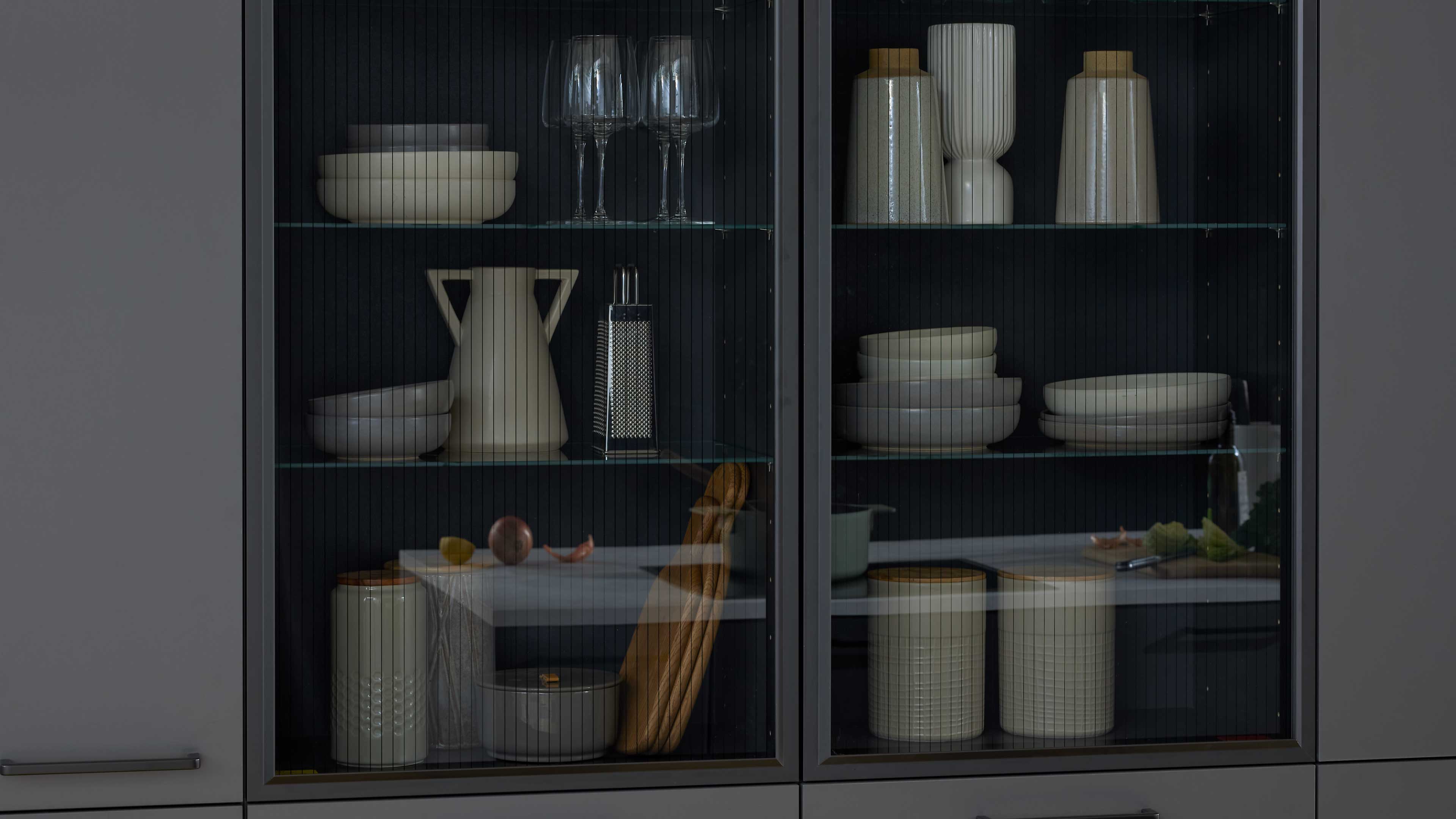 Zweiflügelige Glasschranktür zeigt innen gestapelte weiße Schalen, Weingläser, Vasen, Reibe, Vorratsdosen und ein Baguette.