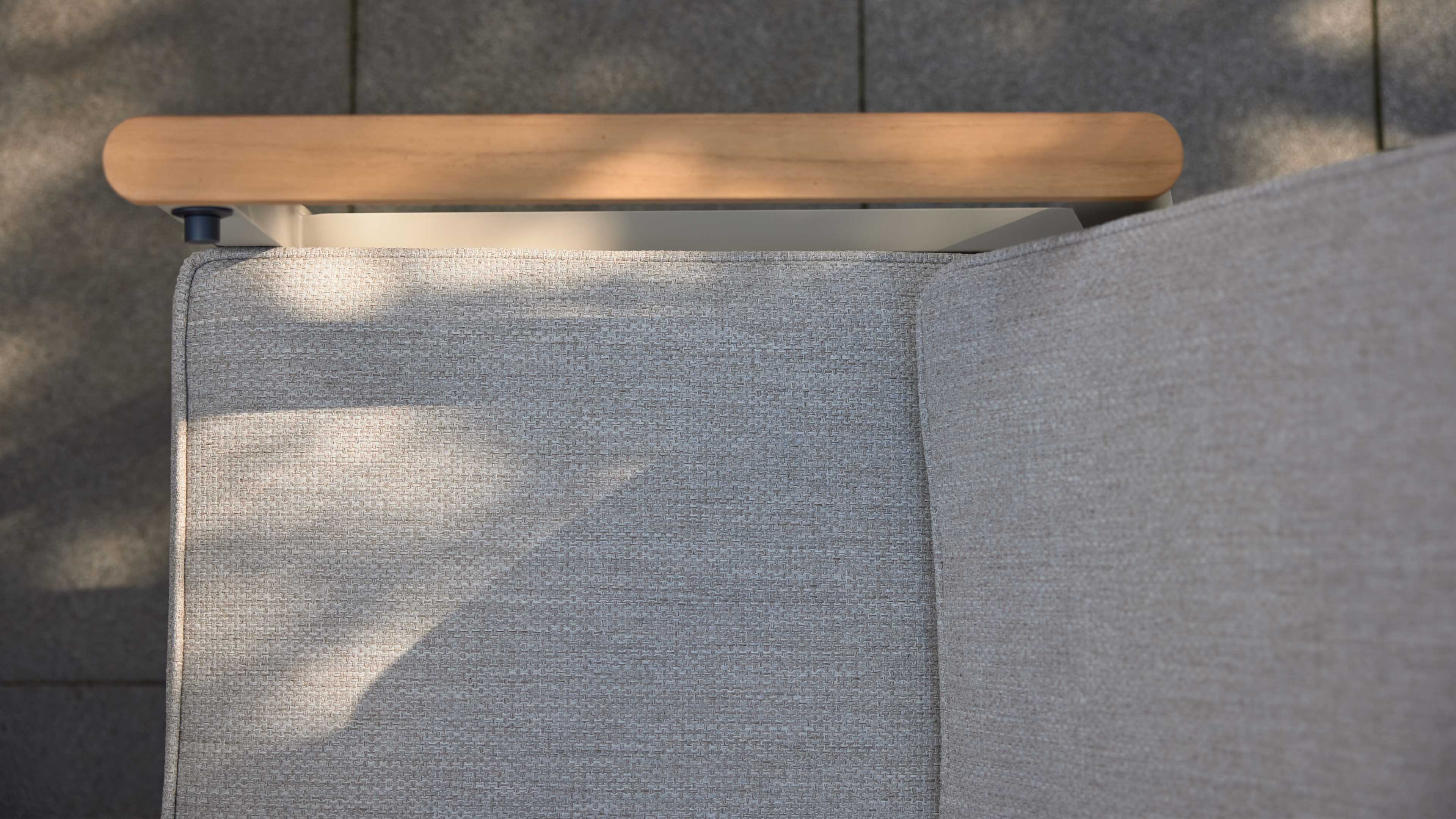 Grauer Sofa-Sitz mit sichtbarer Webstruktur und Rückenlehne, oben ein abgerundetes Holzarmteil und Schatten auf Bodenfliesen.