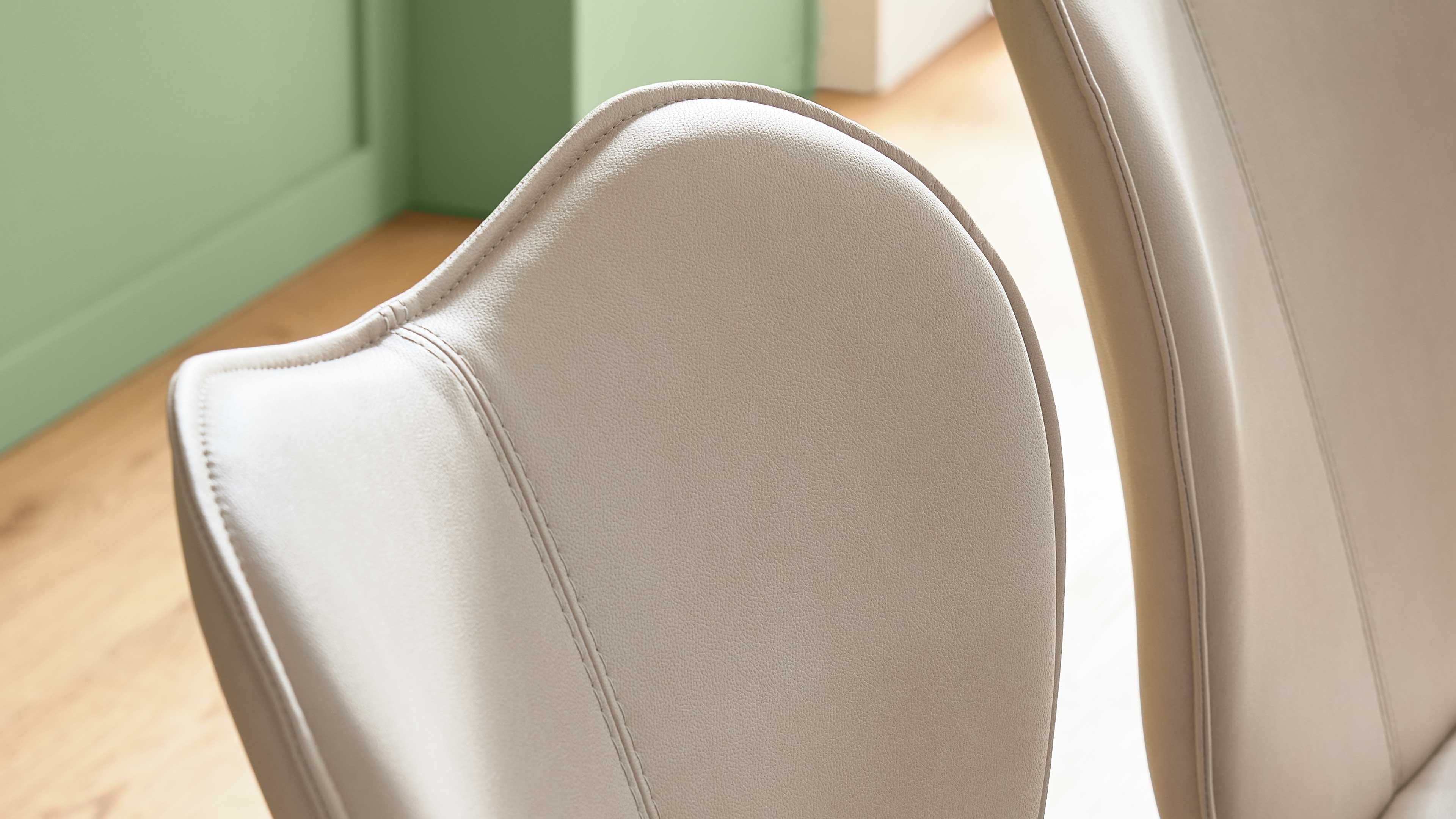 Heller beigefarbener Polsterstuhlrücken mit zwei sichtbaren Nähten, daneben weiterer Stuhlrücken, auf Holzfußboden vor grüner Wand.
