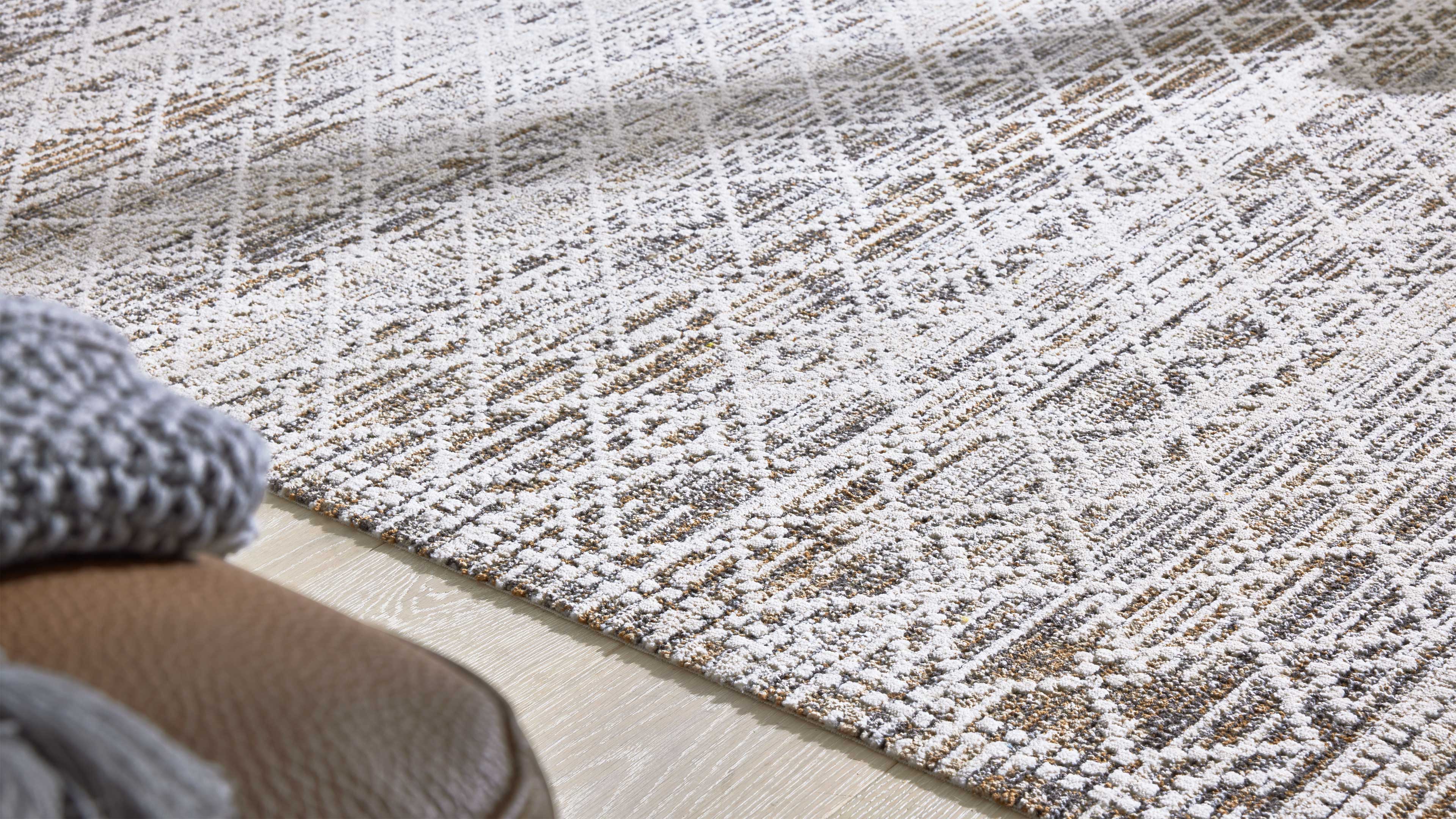 Teppich mit weiß-grau-beigem Rautenmuster auf hellem Holzfußboden, links Lederhocker und graue Strickdecke teils sichtbar.