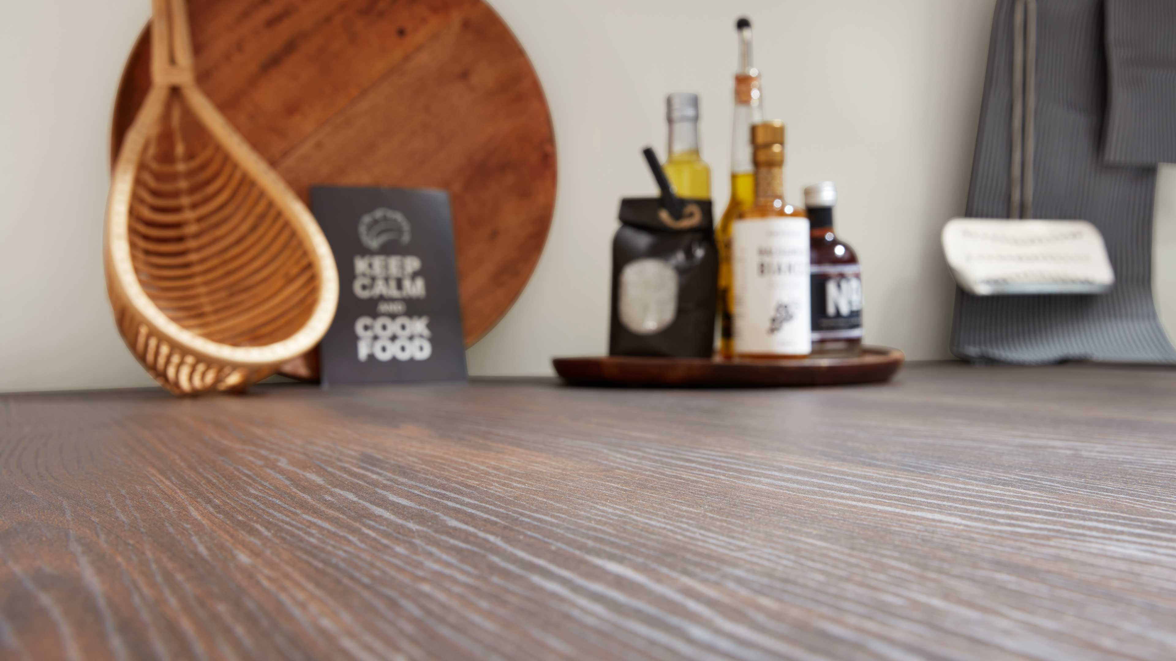 Nahaufnahme einer strukturierten Holzarbeitsplatte, im Hintergrund rundes Schneidebrett, hängender Korb, Schild und Tablett mit Flaschen.