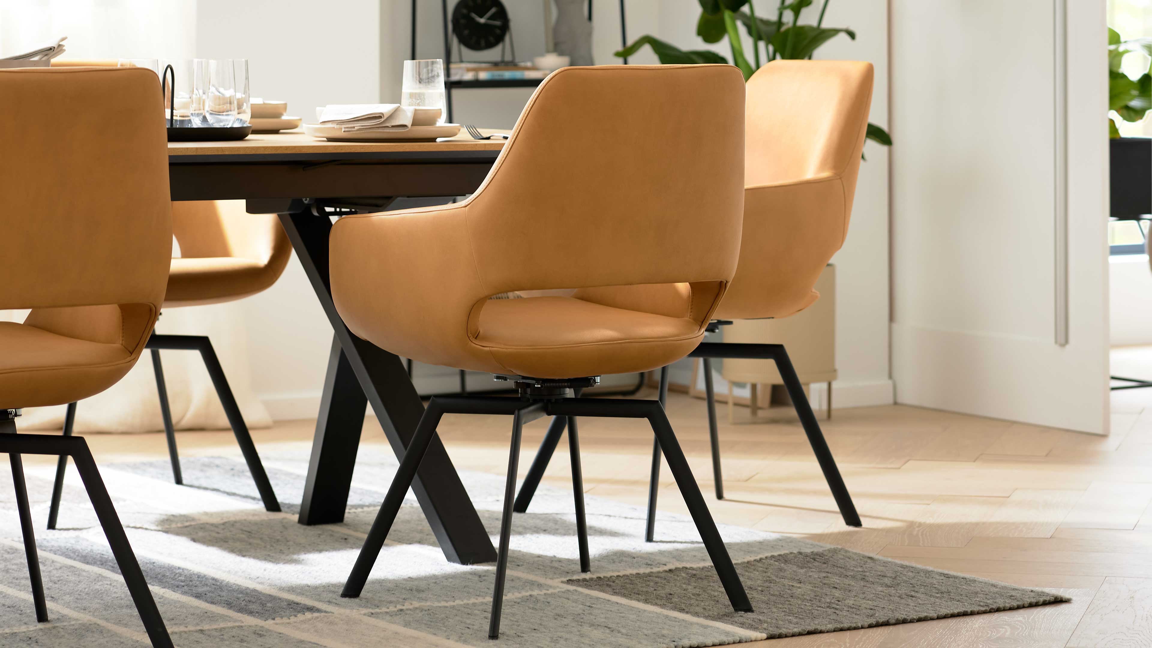 Cognacfarbener Esszimmerstuhl mit offenem unteren Rücken, schwarzem Metallgestell und schrägen Beinen neben gedecktem Holztisch.
