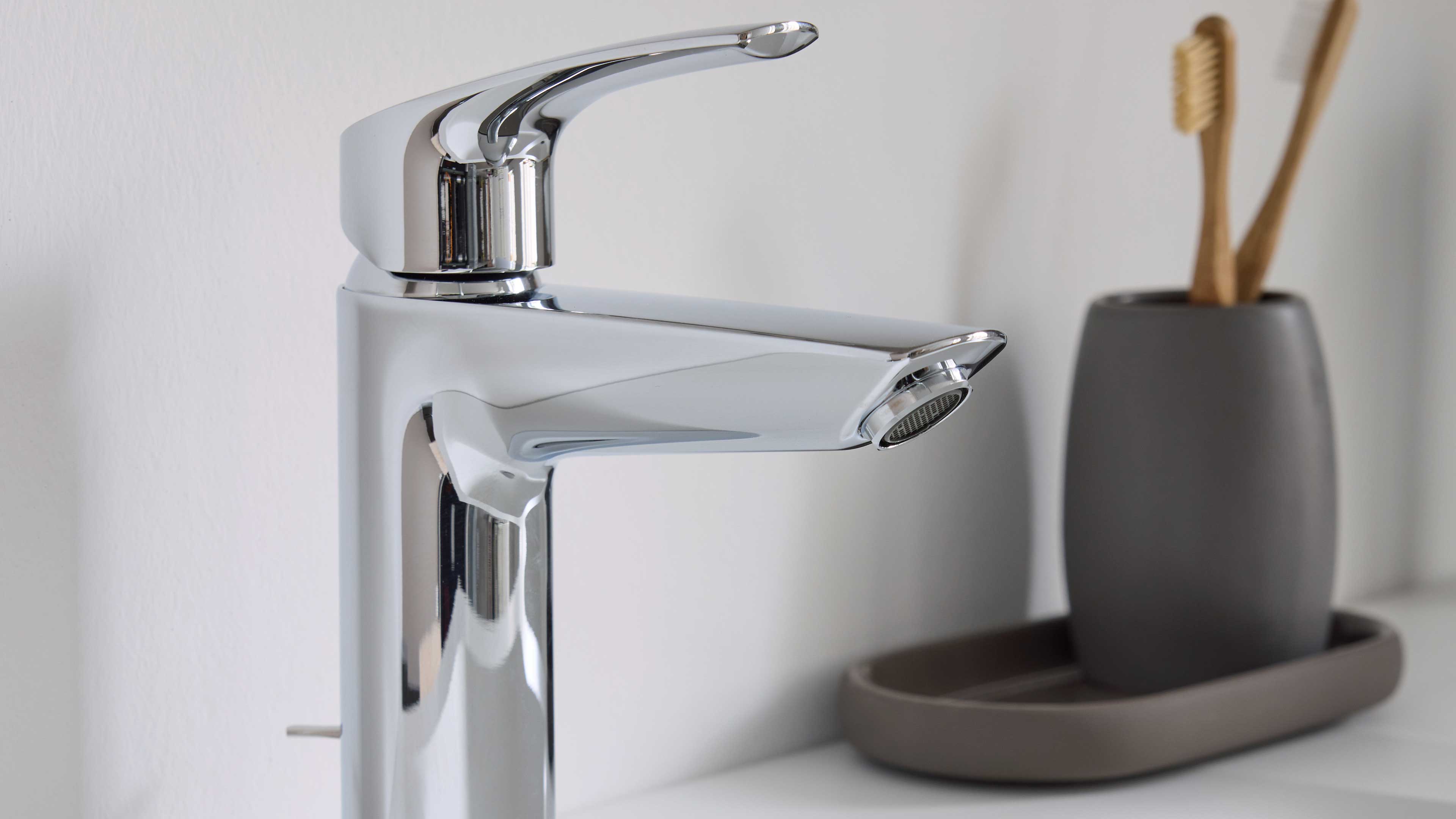 Verchromter Einhebel-Waschtischmischer vorn links, rechts grauer Zahnputzbecher mit zwei hölzernen Zahnbürsten auf Ablage.