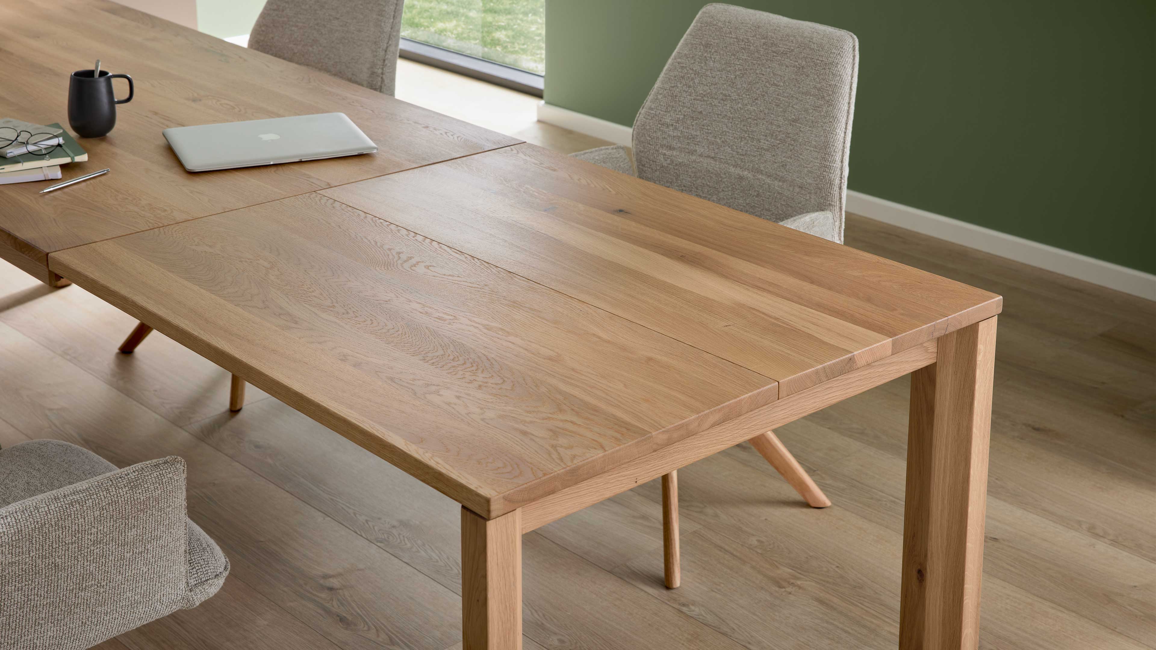 Eichefarbener Tisch mit sichtbarer Fuge, umgeben von hellgrauen Polsterstühlen, Laptop, Becher und Brille.
