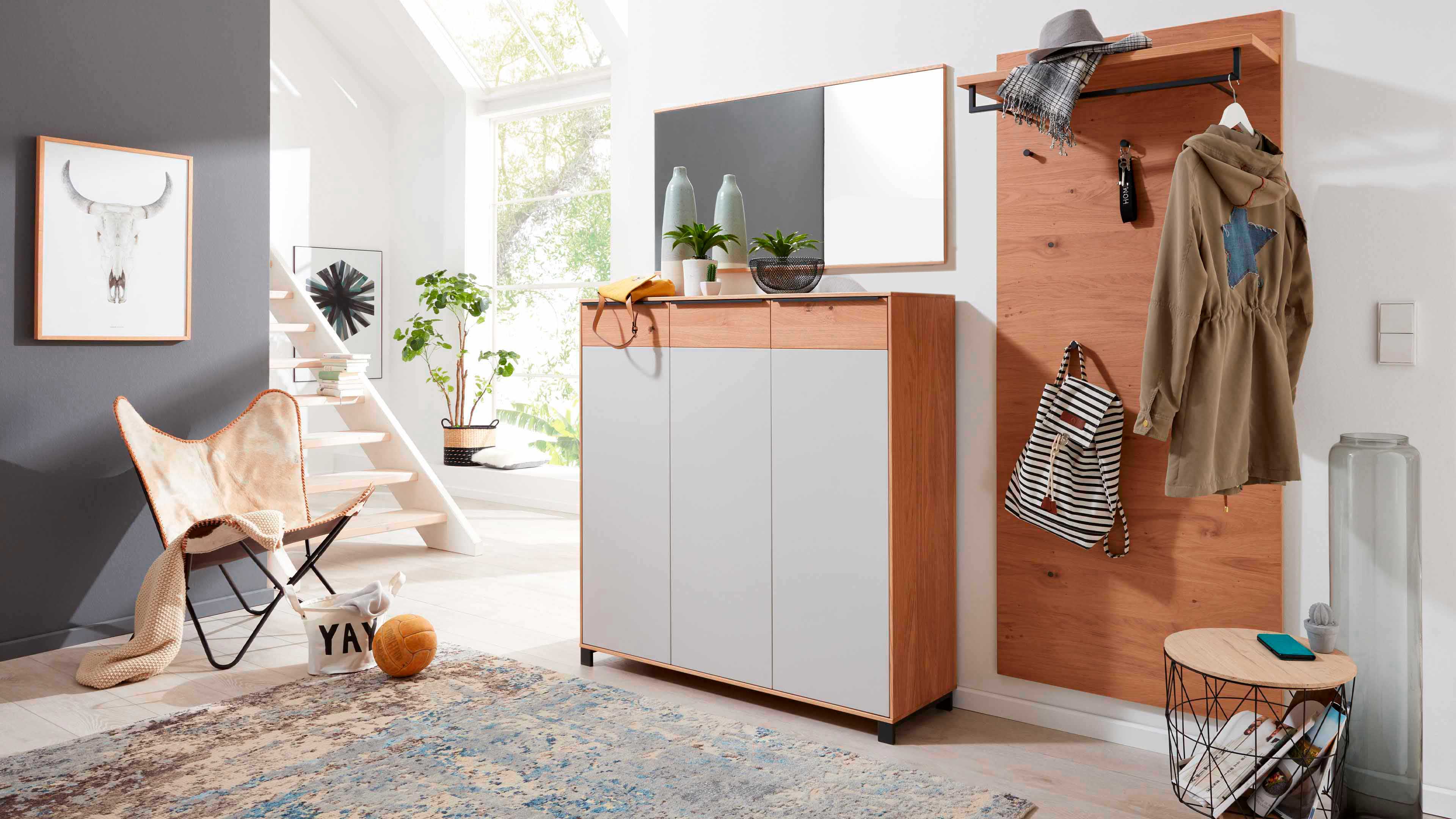 3-türiger Holzschrank mit grauen Fronten und Spiegel, darauf Vasen, Pflanzen; rechts Paneel mit Hut, Schal, Jacke und Rucksack.