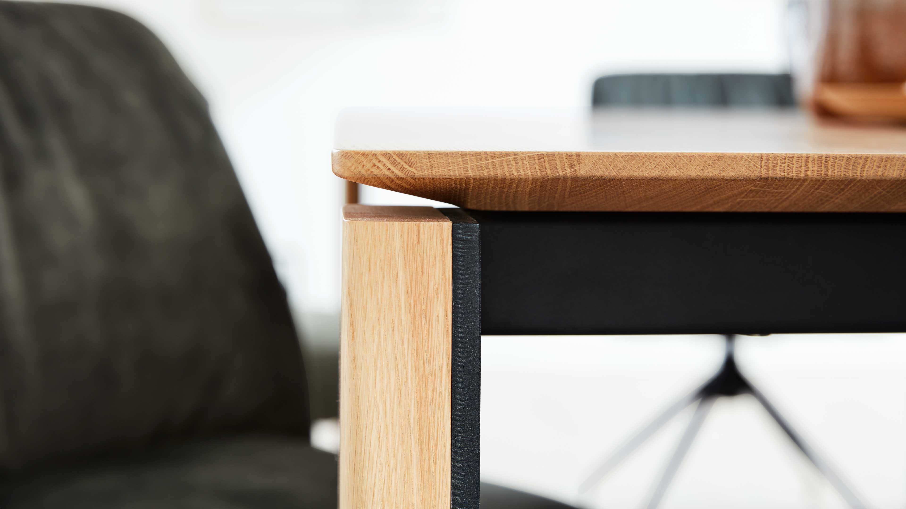Vorderer Tischbereich mit dickem hellen Holzplattenrand und schwarzem Metall-Unterzug, links unscharfer dunkler Stuhl im Hintergrund.