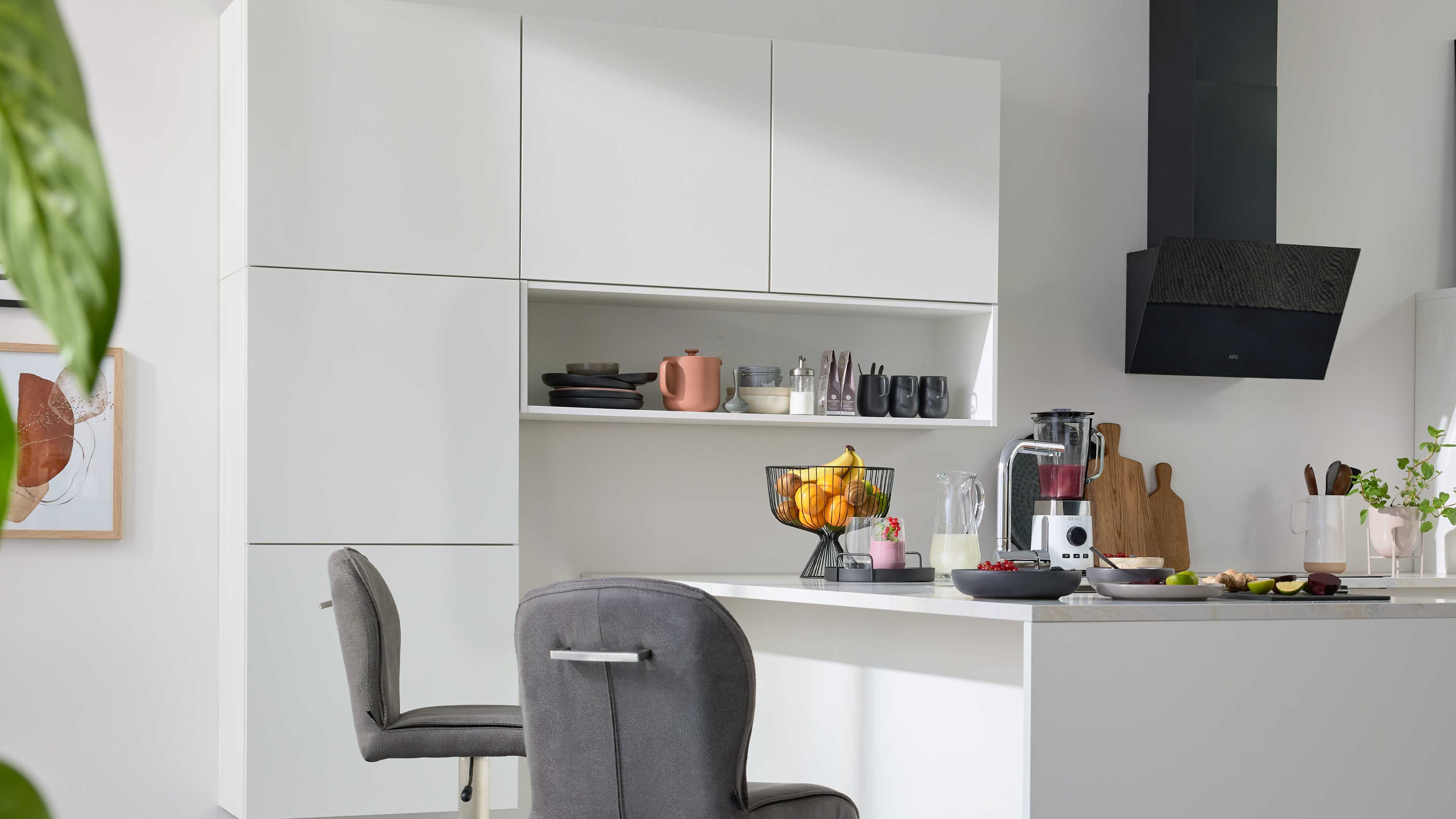 Weiße Kücheninsel mit zwei grauen Barhockern, offener Küchenzeile mit Regalfach, Obstkorb, Mixer und schwarzer Dunstabzugshaube.