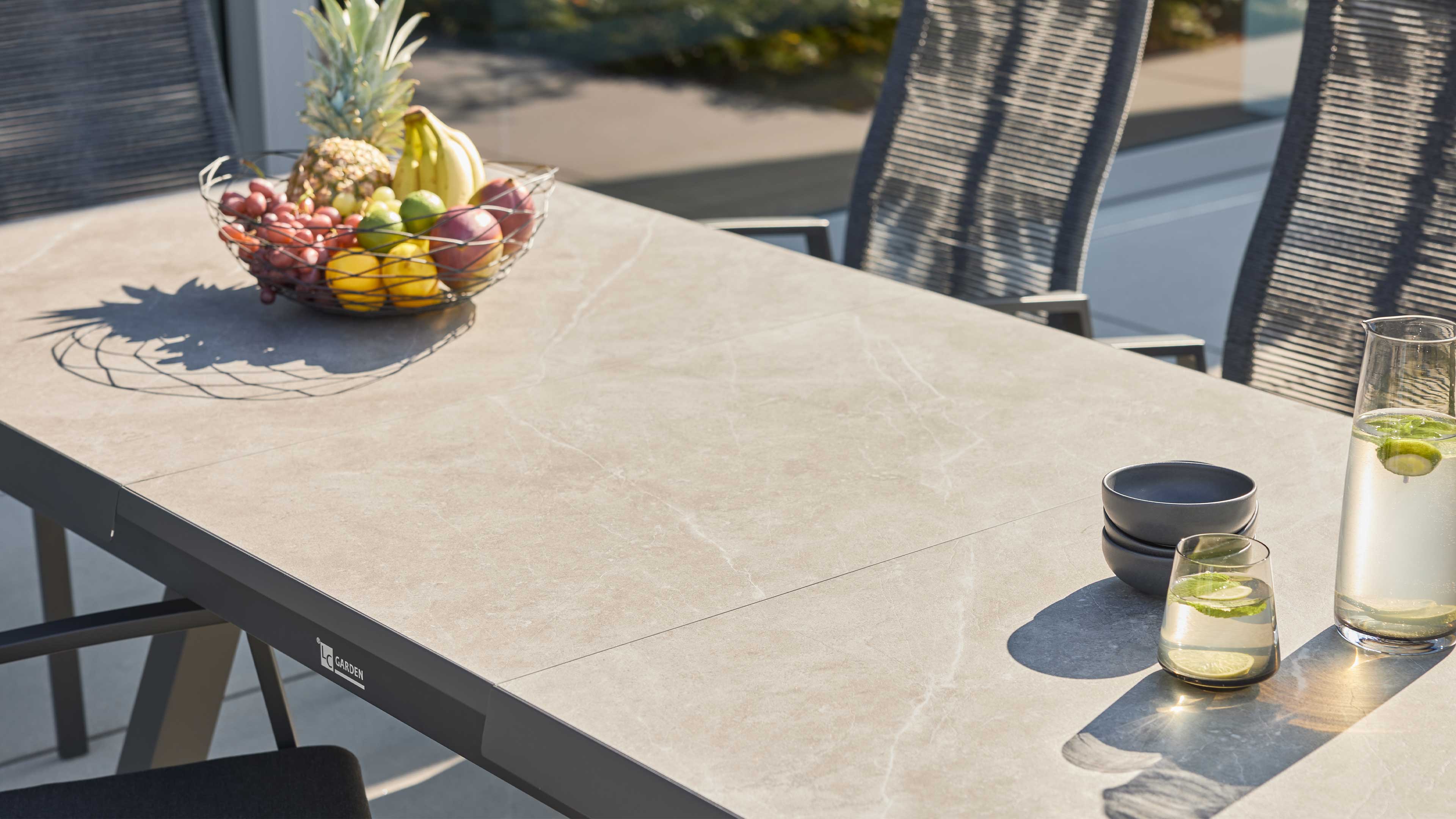 Rechteckiger Terrassentisch mit hellgrauer Oberfläche, links Drahtkorb mit Früchten, rechts Karaffe, Glas und Schalen, Stühle im Hintergrund.